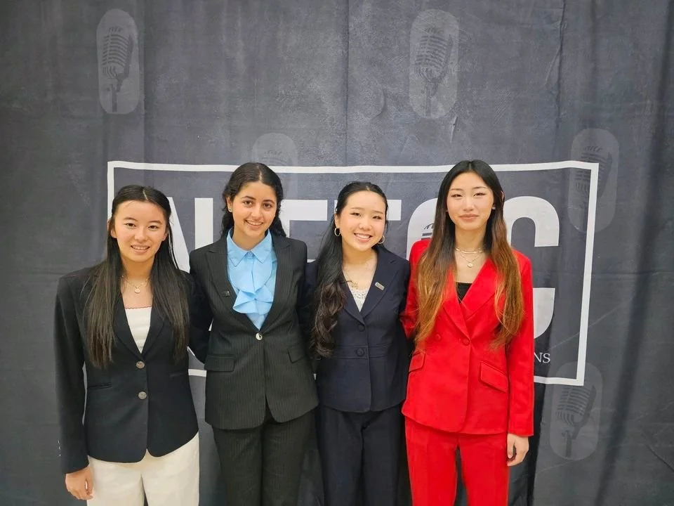 Four women standing side by side in business attire, posing in front of a dark backdrop with the word MIC" and microphone graphics, at a professional event.