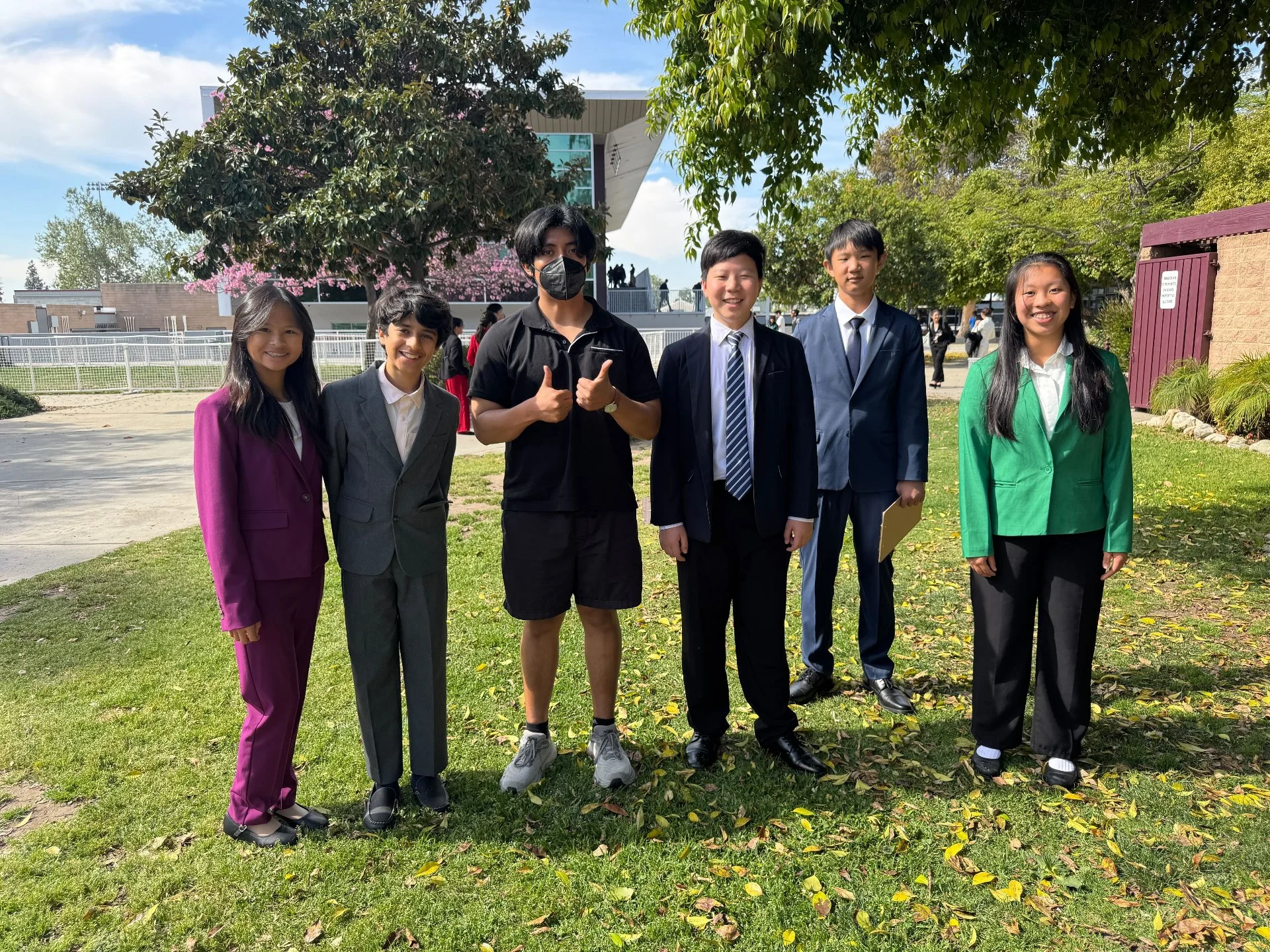 Group of seven students standing outdoors on grass with trees and a modern building in the background.