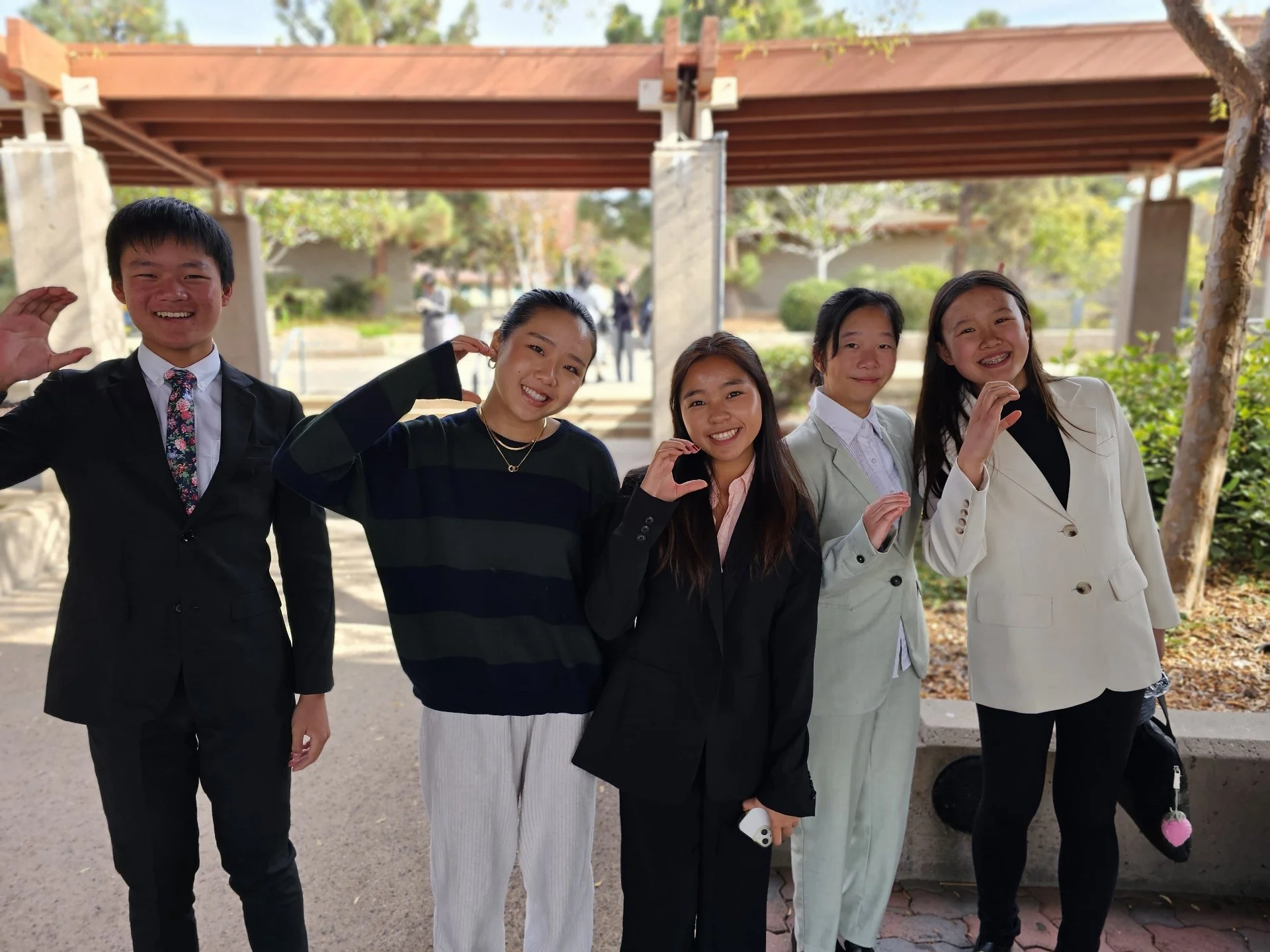 Five young people standing outdoors, smiling and waving at the camera, dressed in semi-formal and casual attire.