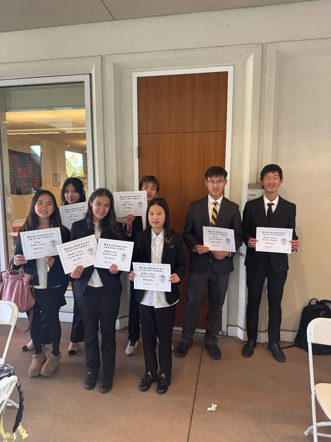 Group of seven students in formal attire holding certificates at an indoor event, standing in front of a wooden door and beige wall.