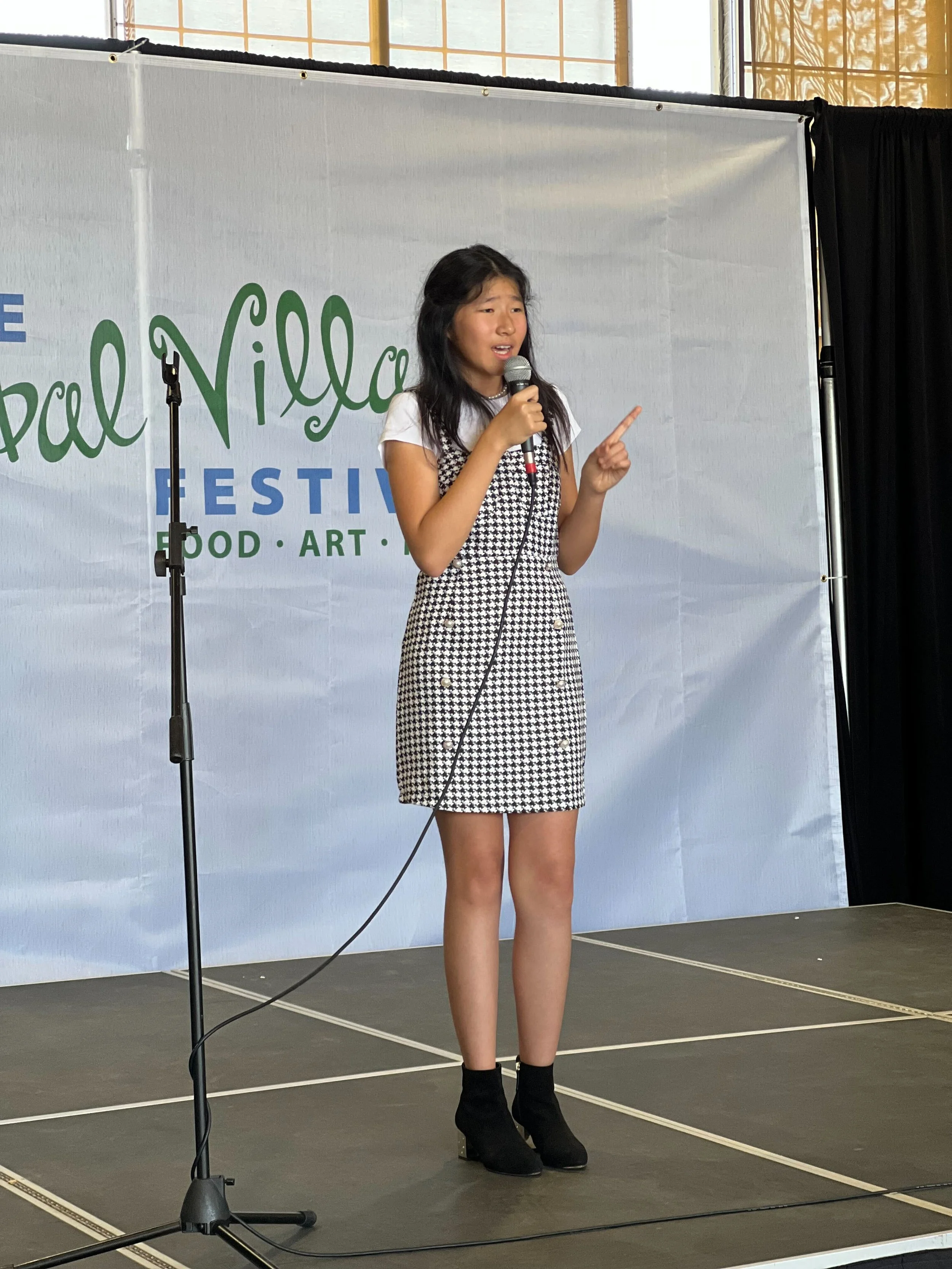 Young girl standing on stage speaking into a microphone at the Pal Valley Festival, wearing a black and white dress and black ankle boots.