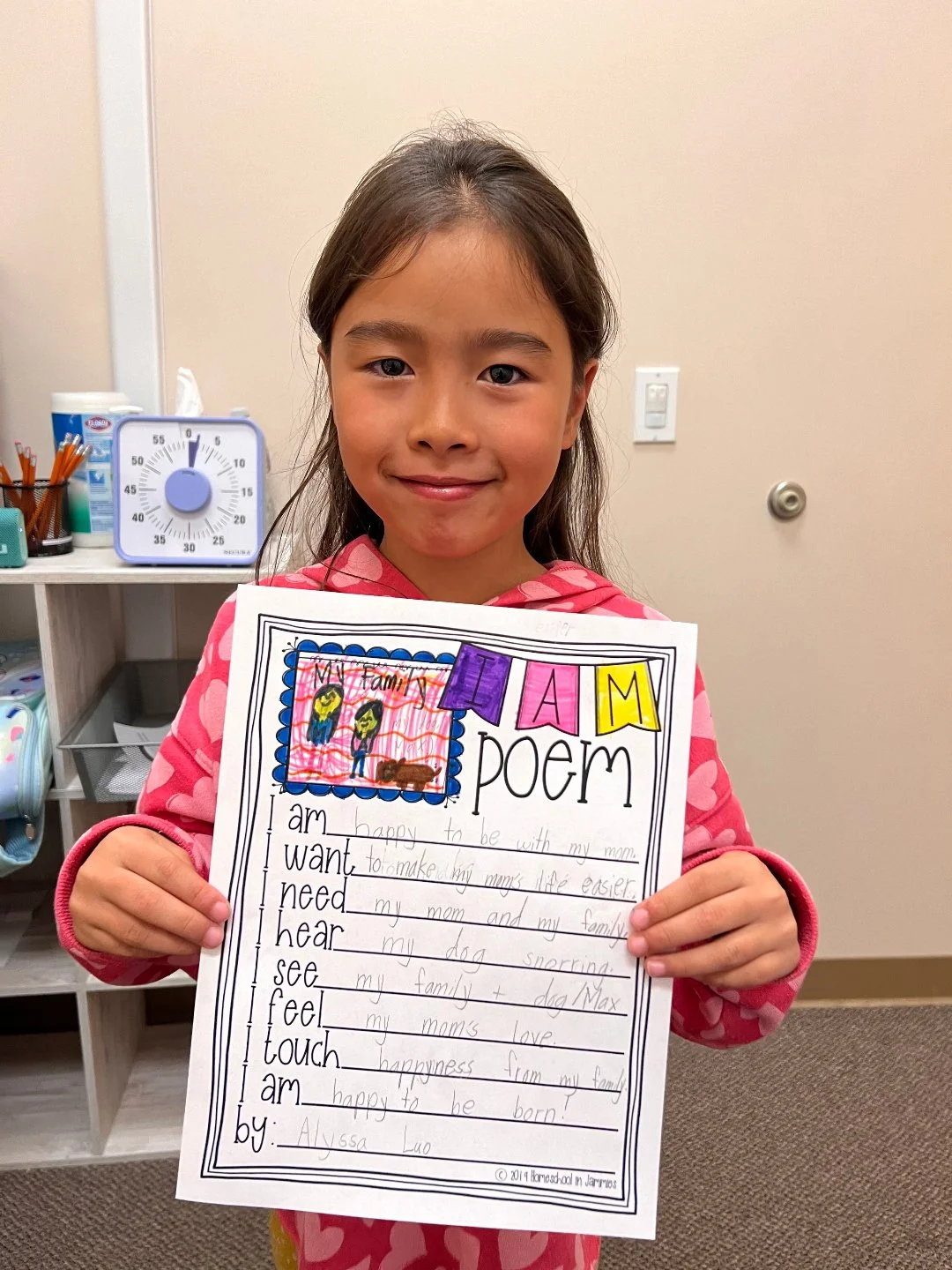 A young girl in a pink patterned hoodie holding a handwritten poem on lined paper with colored decorations and drawings, smiling in a room with shelves and a timer in the background.