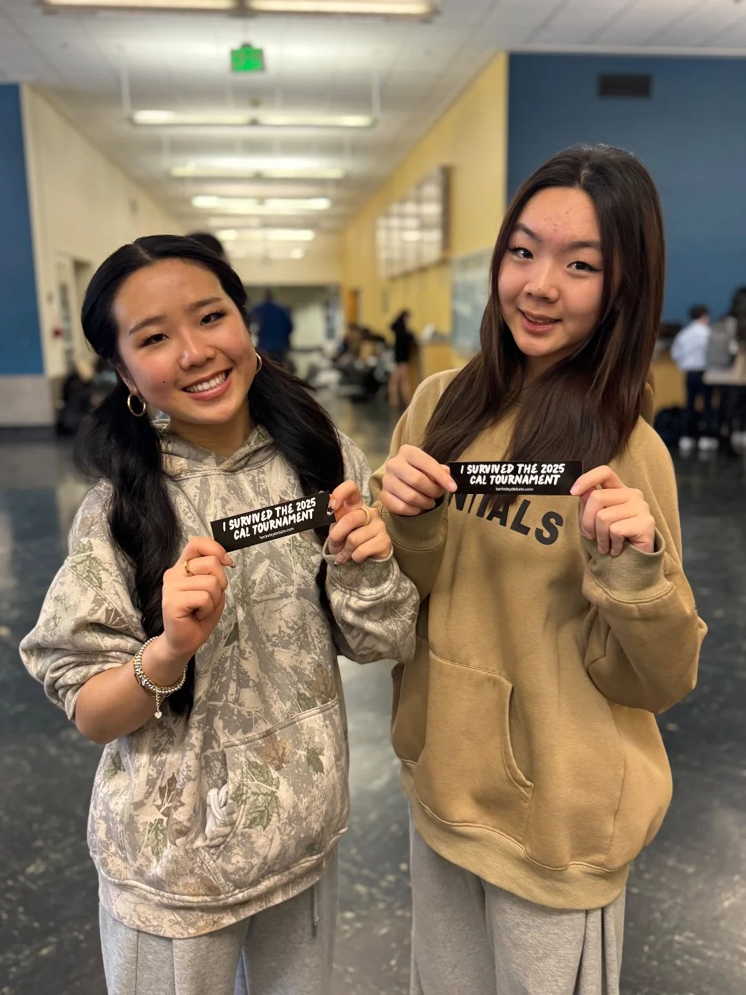 Two young women smiling and holding signs that say 'I survived the 2025 CAL Tournament' inside a school or gymnasium.