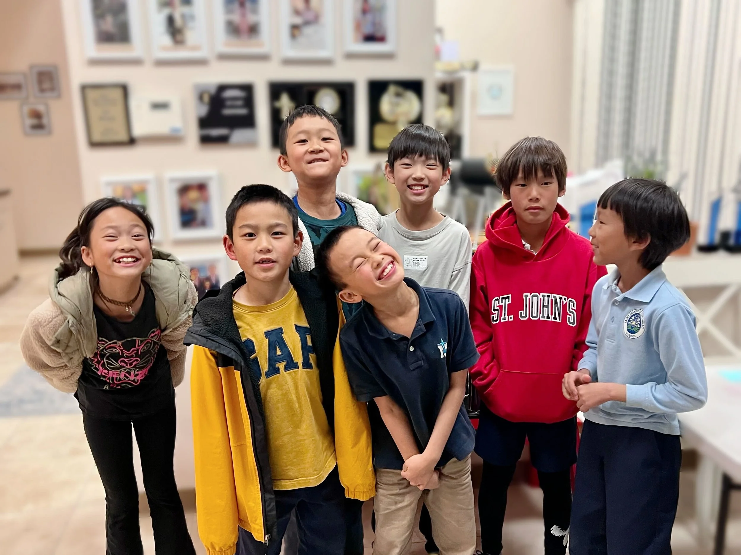 Group of seven children smiling and posing together indoors, casual clothing, standing in front of a wall decorated with framed pictures and awards.