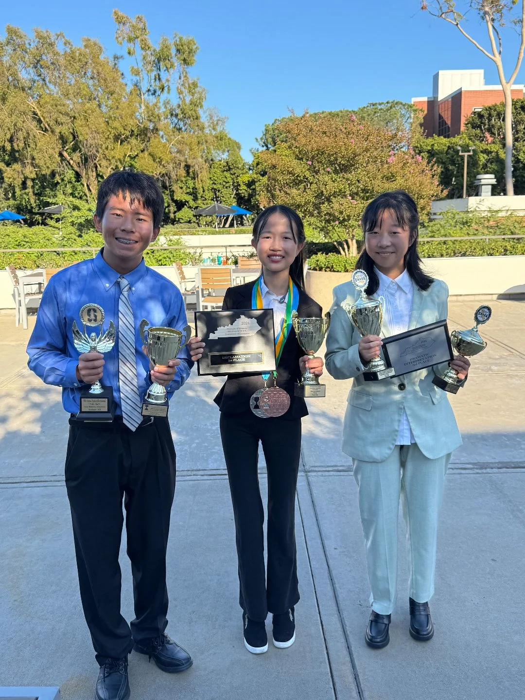 Three children standing outdoors, each holding a trophy and a plaque, dressed in formal attire, with medals around their necks, smiling after an award ceremony.