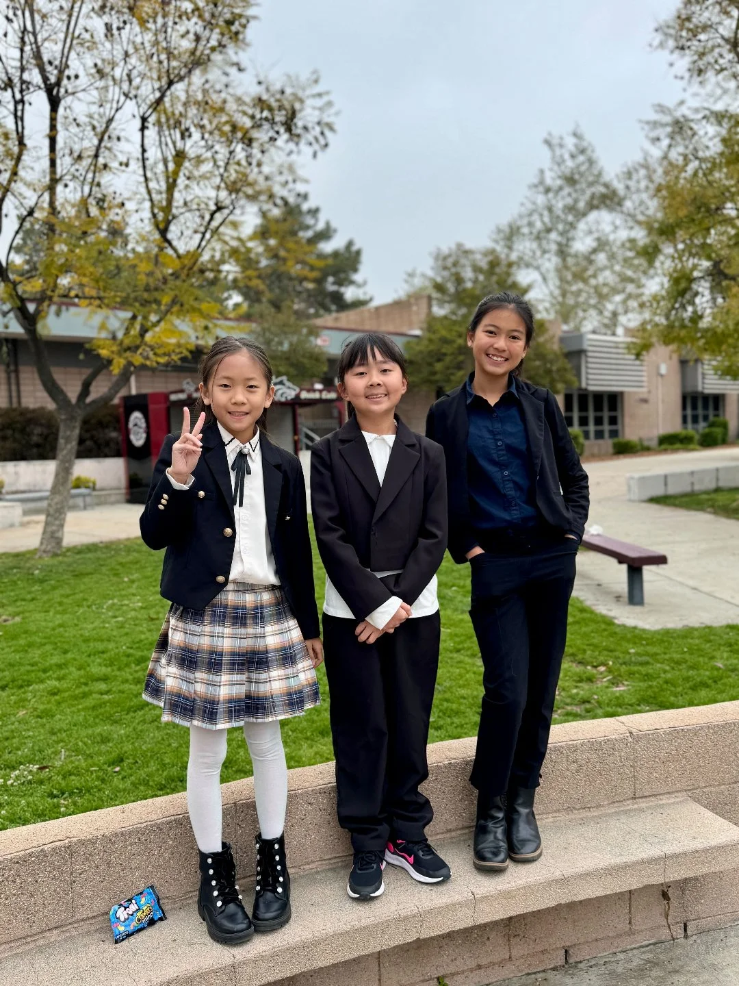 Three young girls standing outdoors on a sidewalk in front of a grassy area with trees and buildings, smiling at the camera. The girl on the left is making a peace sign with her fingers, wearing a dark blazer, white shirt, plaid skirt, white tights, 