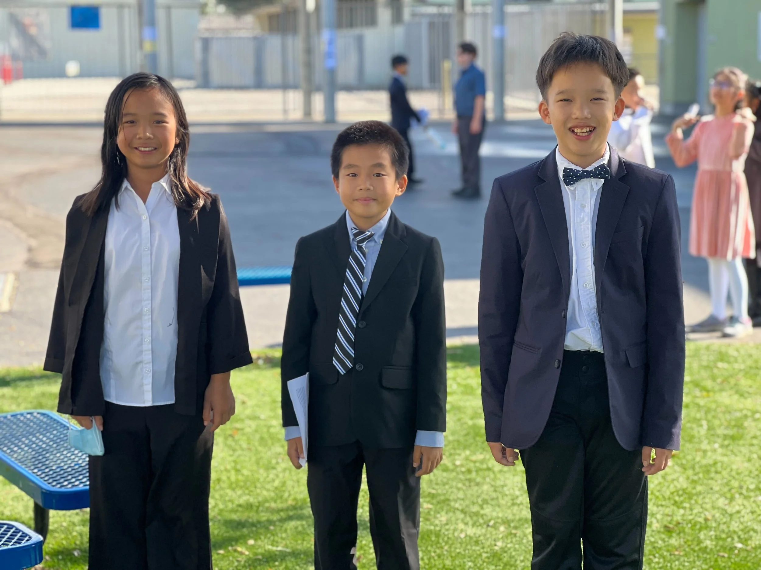 Three children dressed in formal attire standing outdoors on a green lawn, smiling at the camera. The girl on the left holds a face mask, the boy in the middle holds a piece of paper, and the boy on the right wears a bow tie.