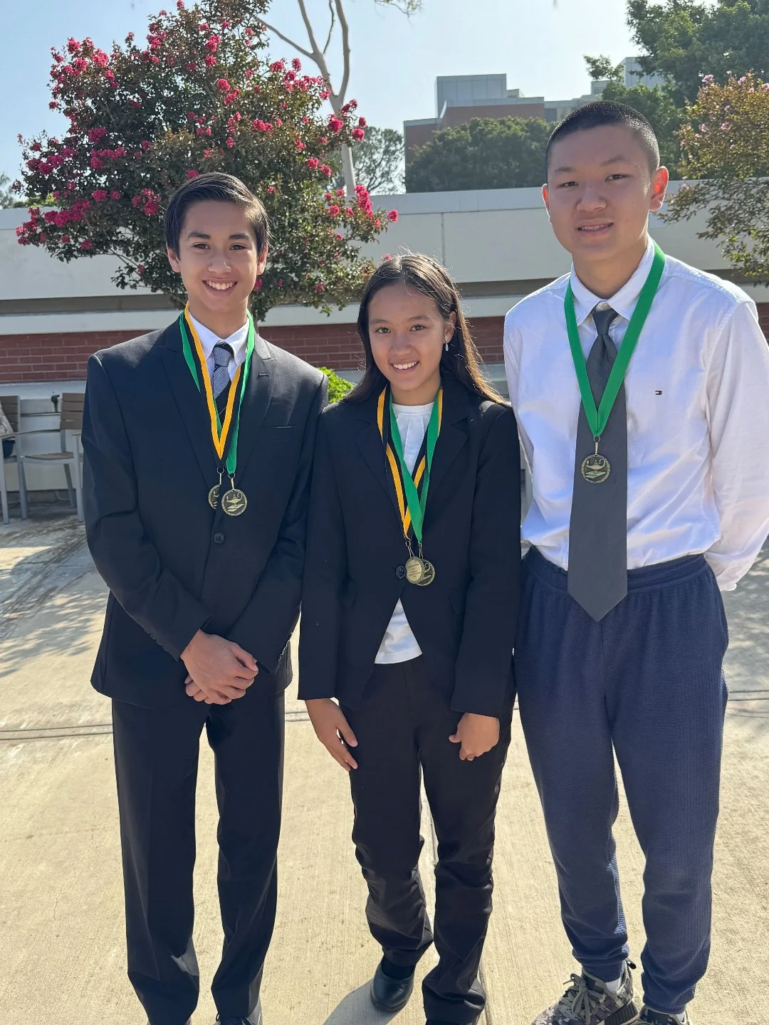 Three teenagers wearing formal attire with medals around their necks, standing outdoors with a flowering tree and modern buildings in the background.