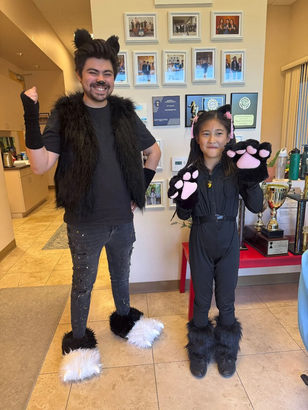A man and a girl dressed in black with cat paw costumes, posing inside a home with framed photos and awards on the wall behind them.