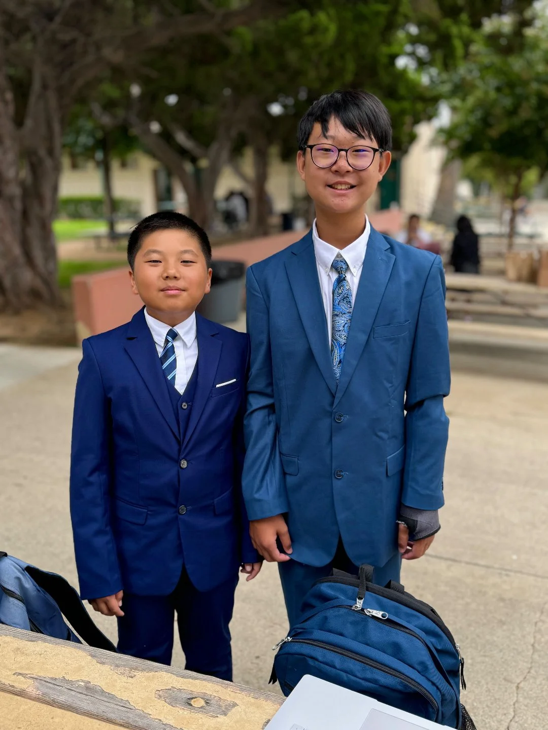 Two boys in blue suits and ties standing outdoors with backpacks, smiling at the camera.