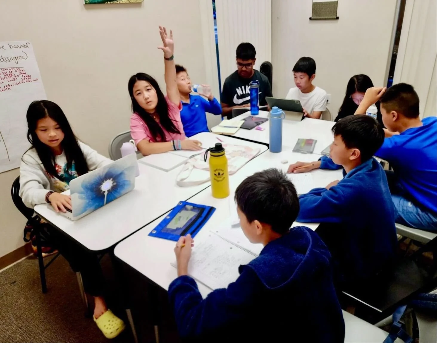 Group of children sitting around a table in a classroom, one girl with her hand raised, some using laptops or tablets, drinking water, and taking notes.