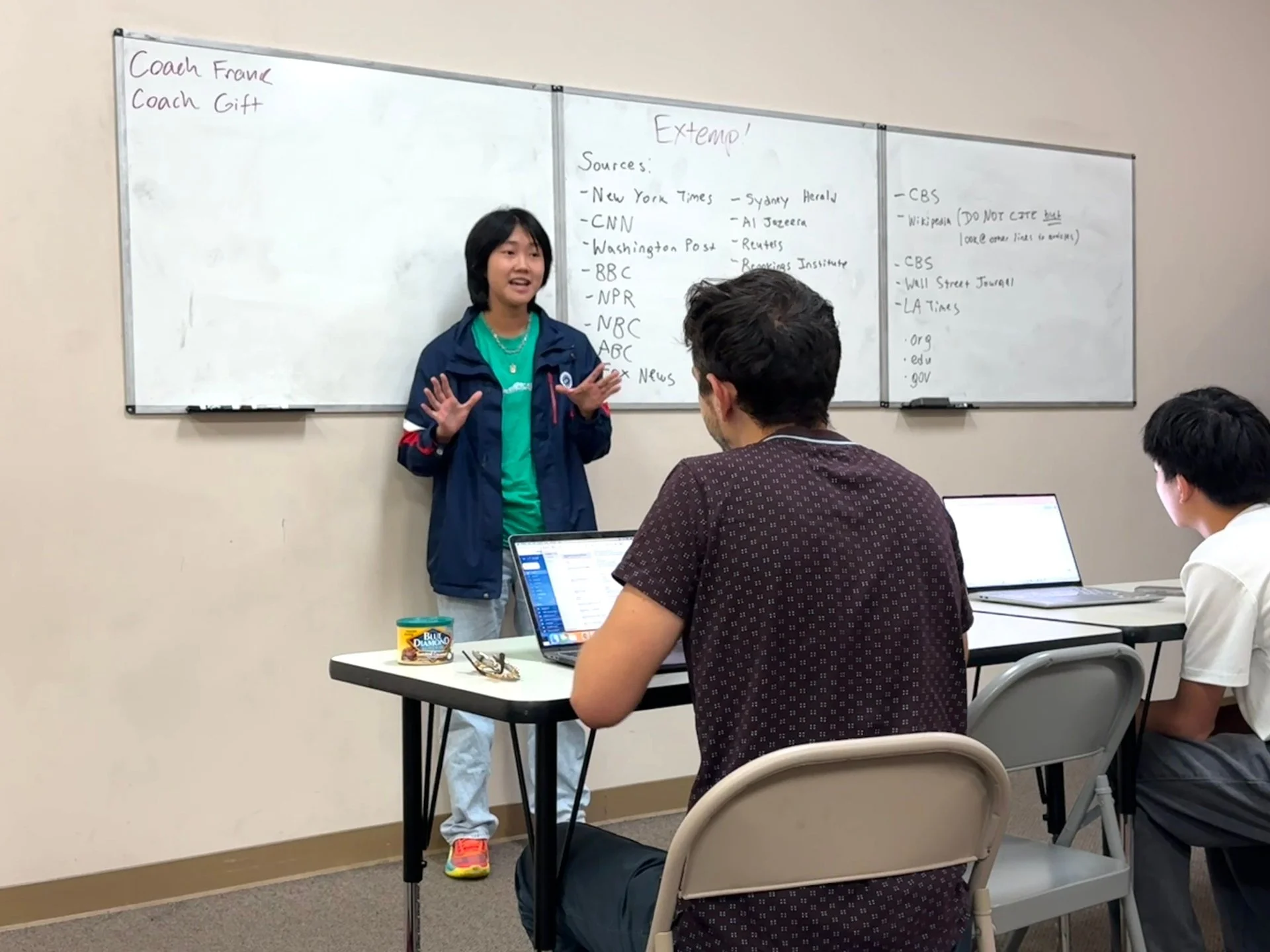 A woman standing and speaking to two seated men in a classroom, with whiteboards behind her filled with notes and sources.
