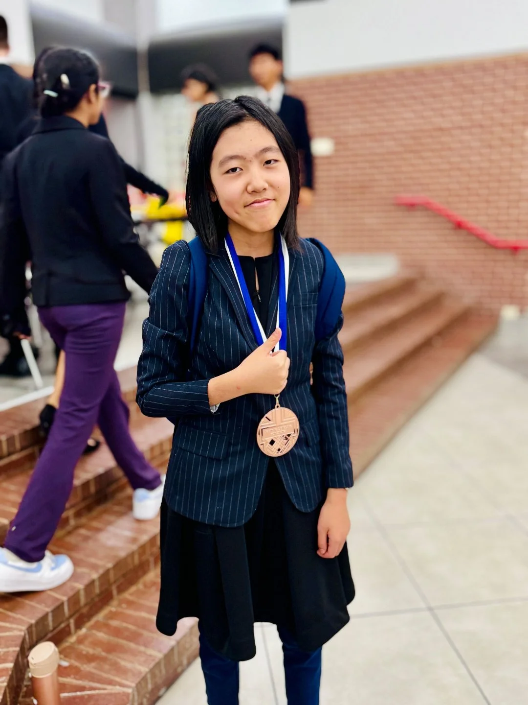 A young girl in a school uniform with a bronze medal around her neck, standing on brick steps at an indoor event.