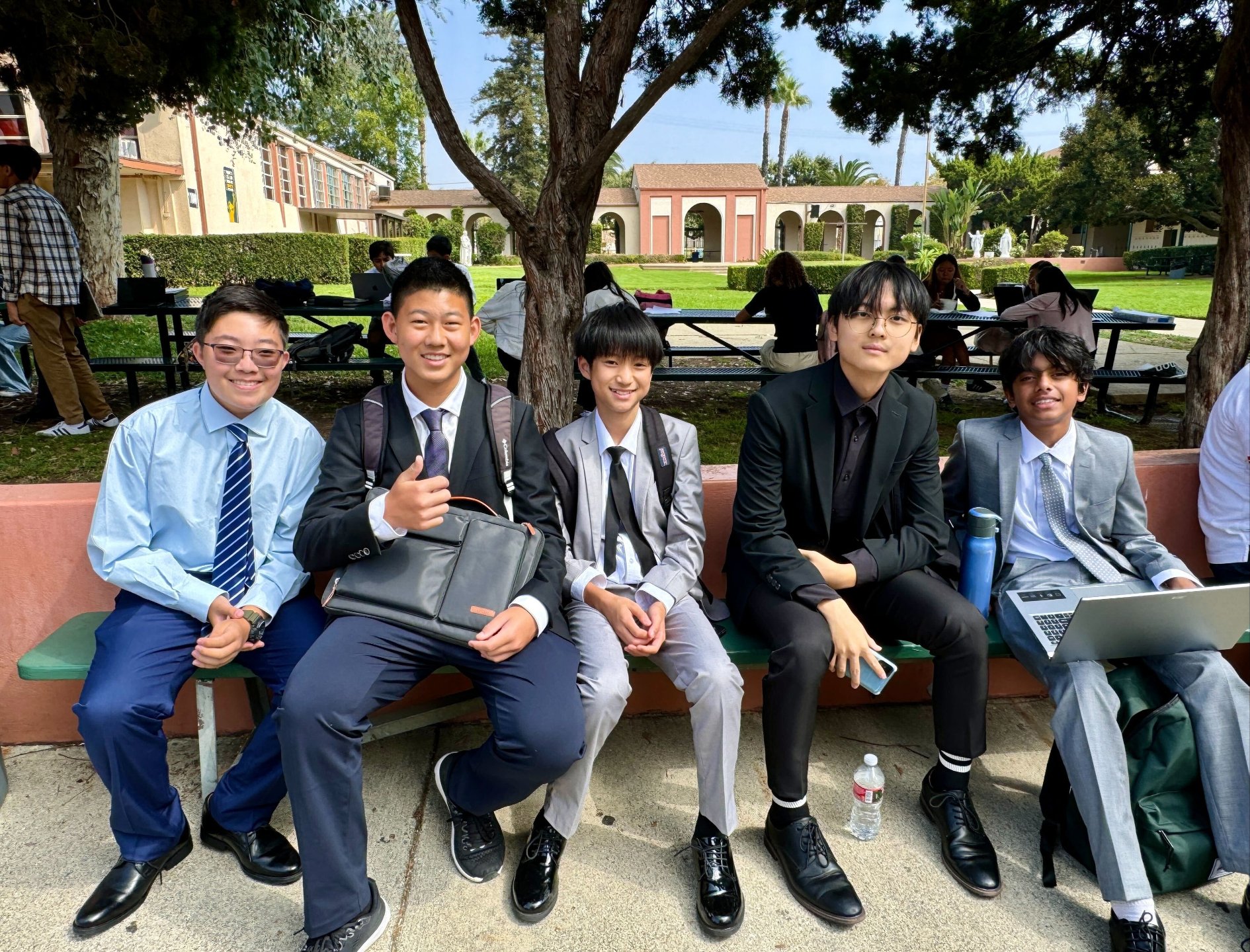 Five boys in suits and school uniforms sitting on a bench outdoors.