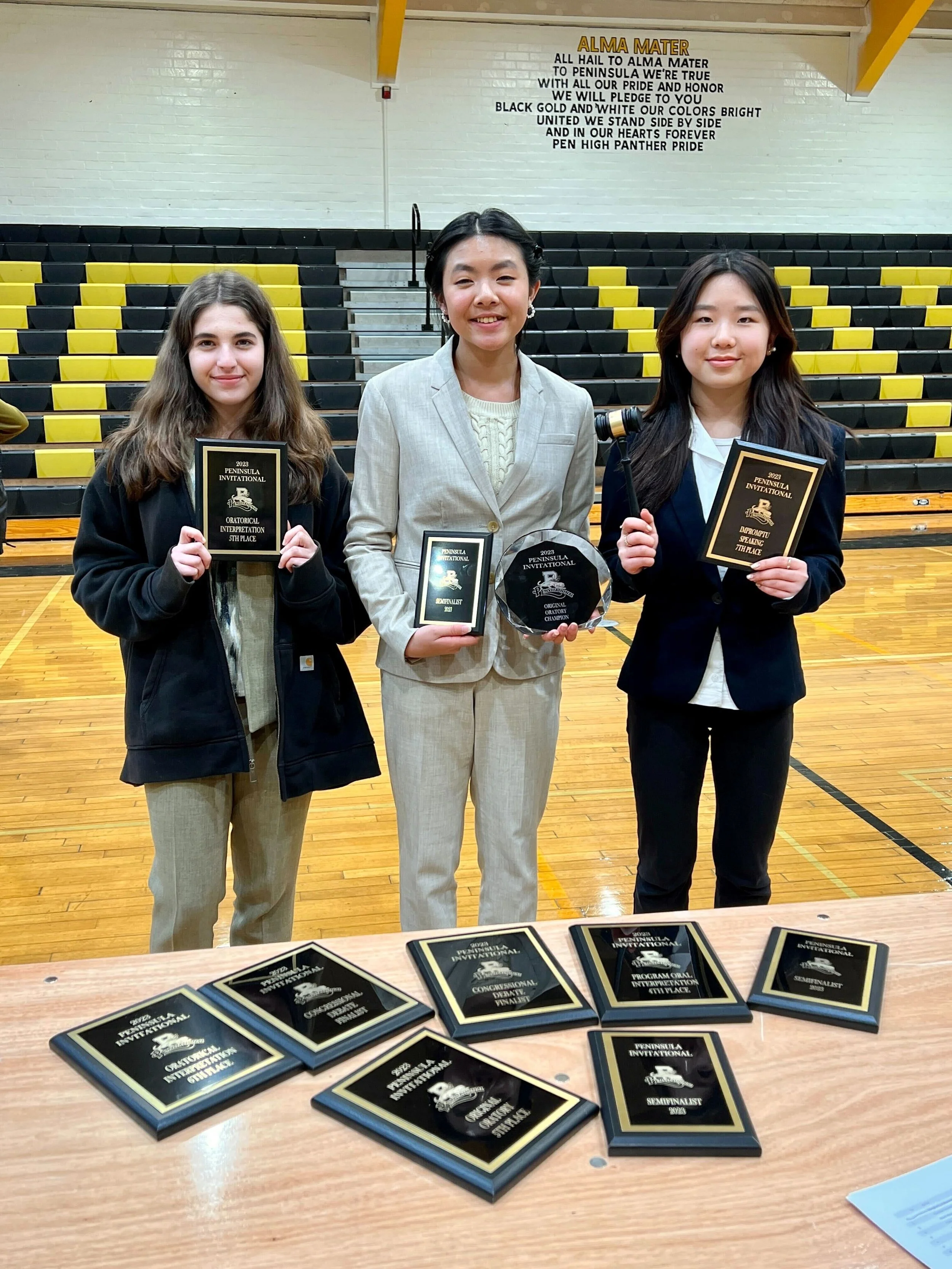 Three young women standing in a gymnasium holding plaques and awards, with a table displaying more plaques in front of them, and a large sign with the word "ALMA MATER" on the wall behind them.
