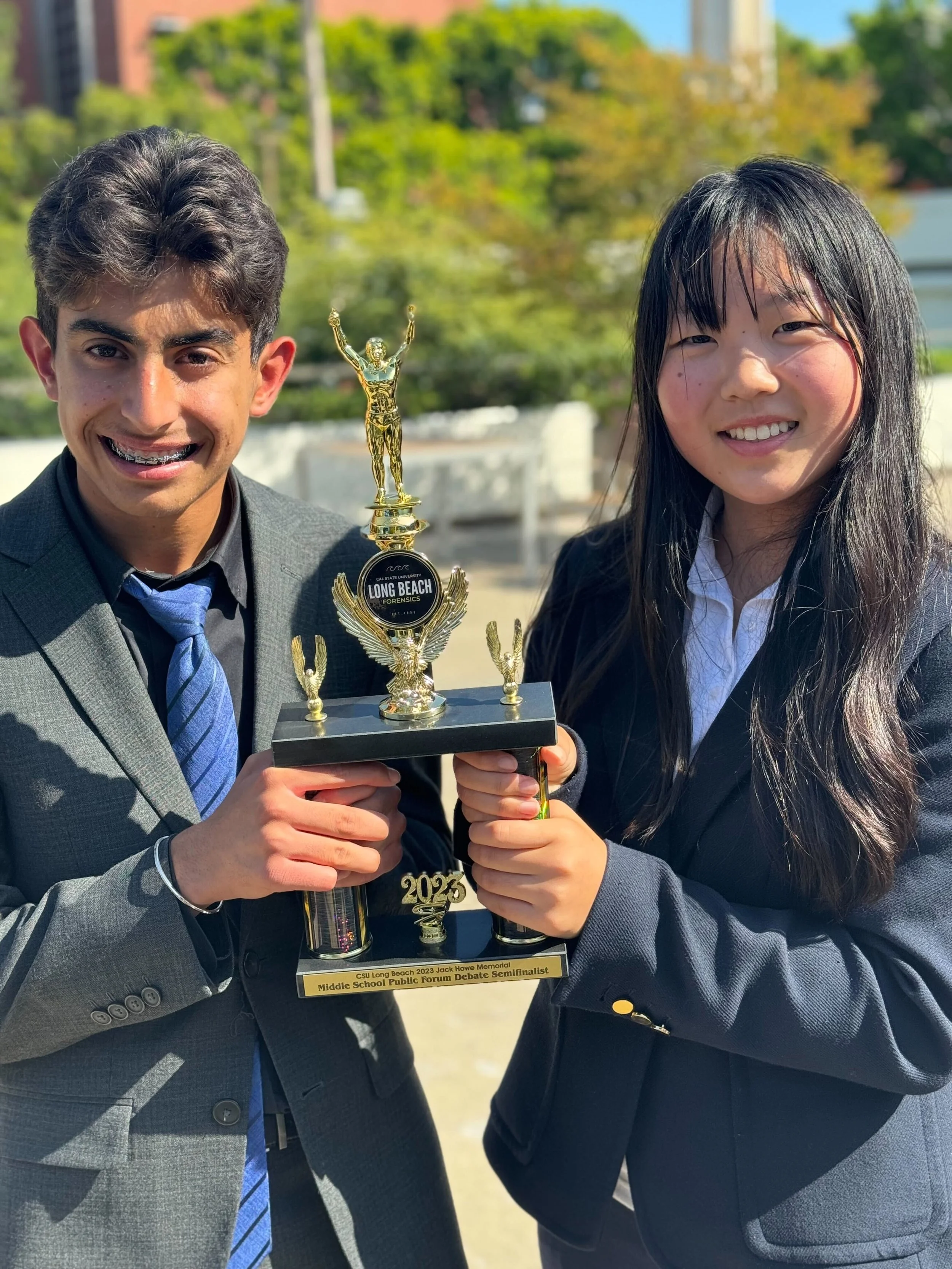 Two students in business attire holding a trophy with the label 'CSU Long Beach 2023 Jack Howe Memorial Middle School Public Forum Debate Semifinalist' outdoors on a sunny day.