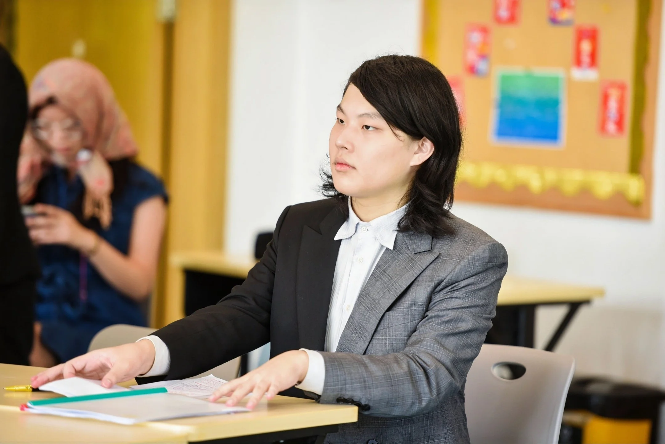 A young woman in a business suit sitting at a desk in a classroom or office, focusing on paperwork or a task.