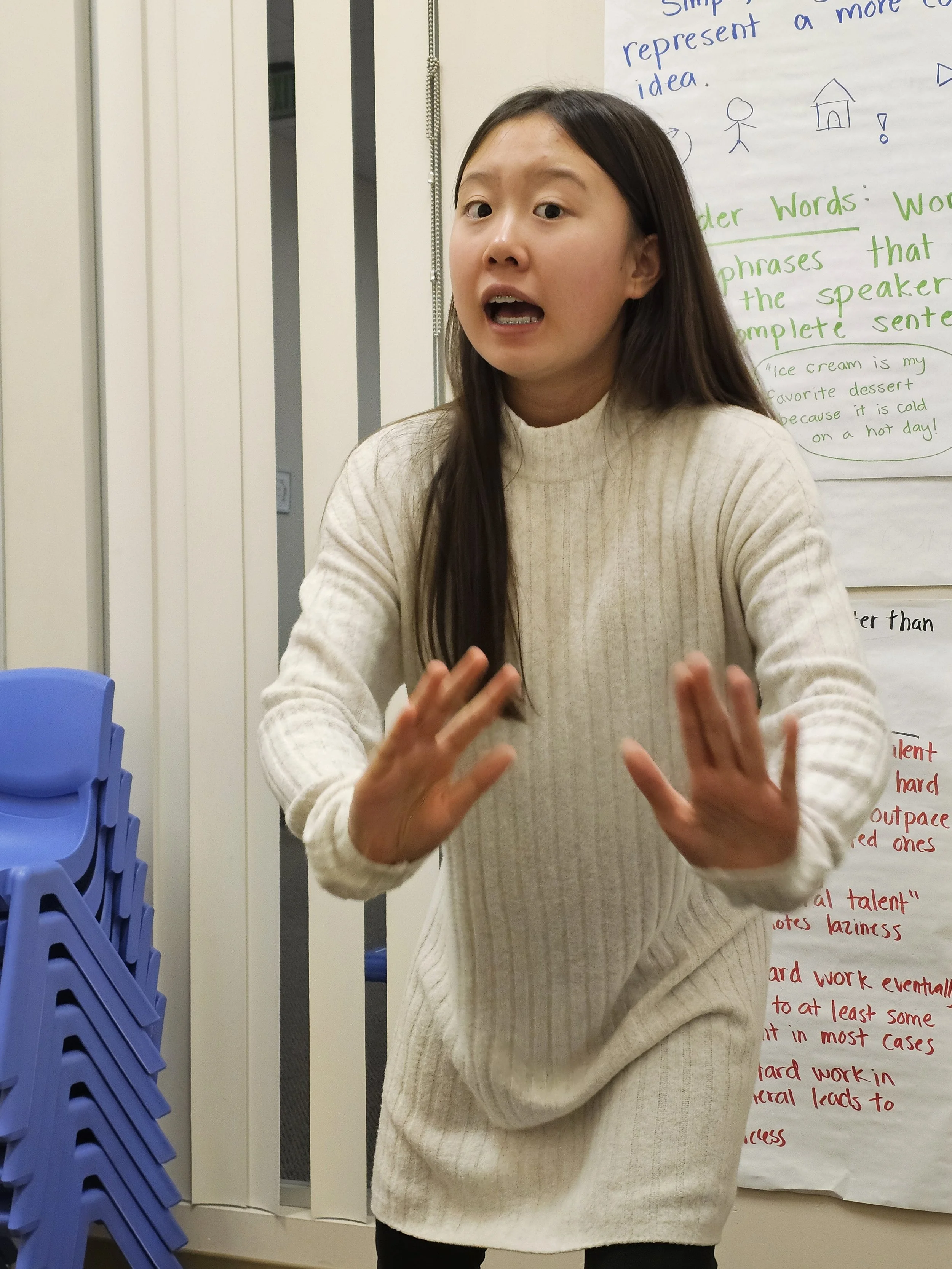 A woman with long dark hair wearing a white ribbed sweater, speaking and gesturing with her hands in front of a whiteboard with handwritten notes.