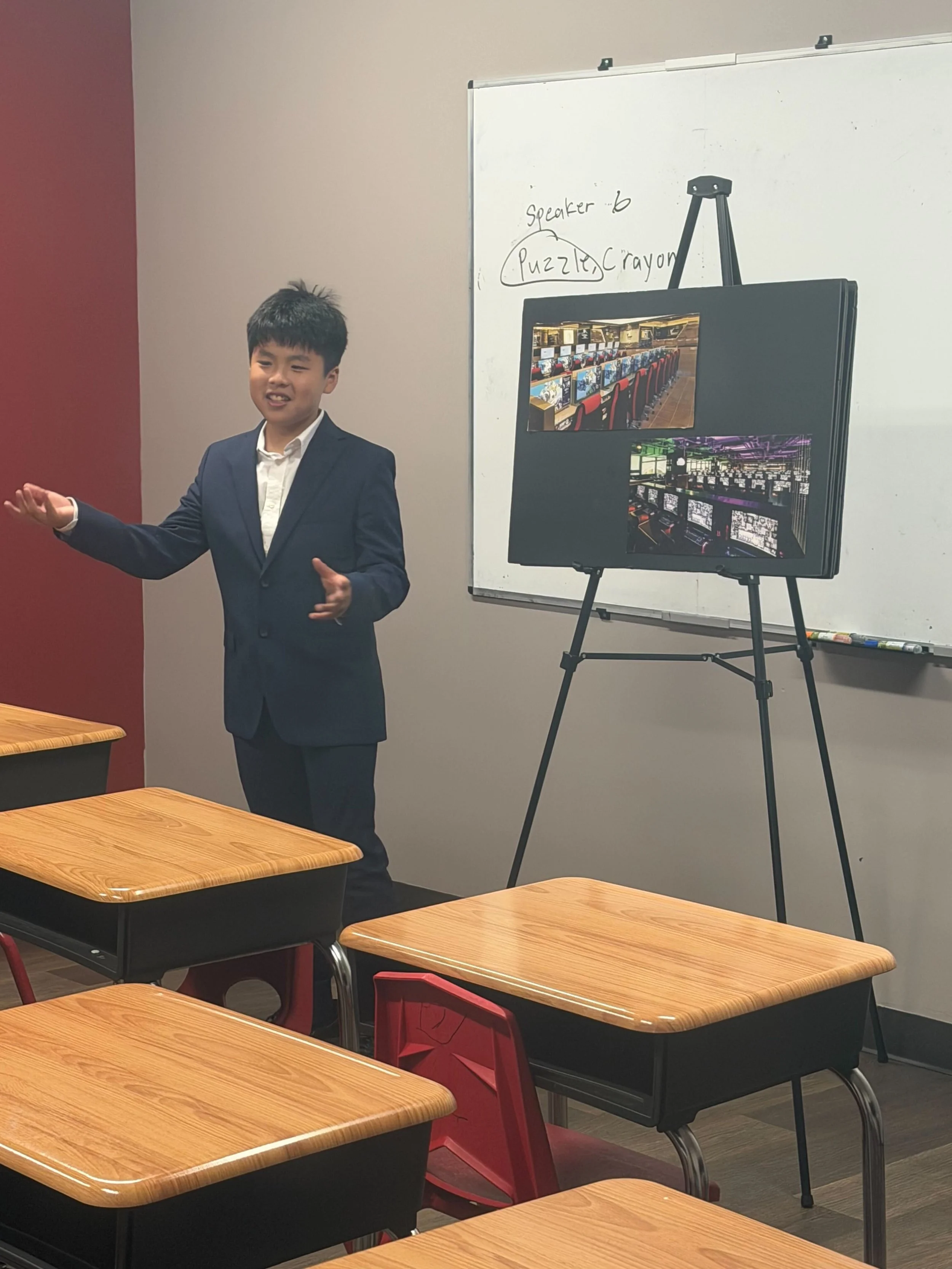 A young boy in a navy suit giving a presentation in a classroom. There are empty desks and a whiteboard in the background with writing that says 'speaker' and 'puzzle, crayon.' A standing display board shows images of a game arcade and a computer lab