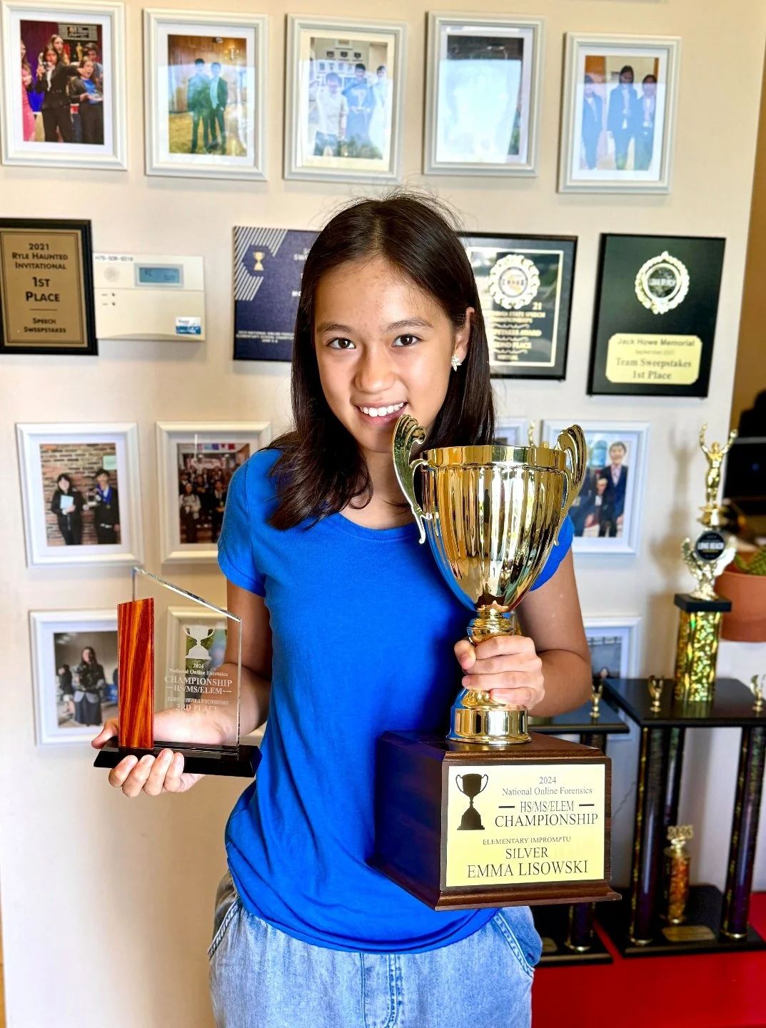 A young girl in a blue shirt holding a large gold trophy and a smaller clear, red, and wood-colored award, standing in front of a wall displaying framed pictures and plaques.