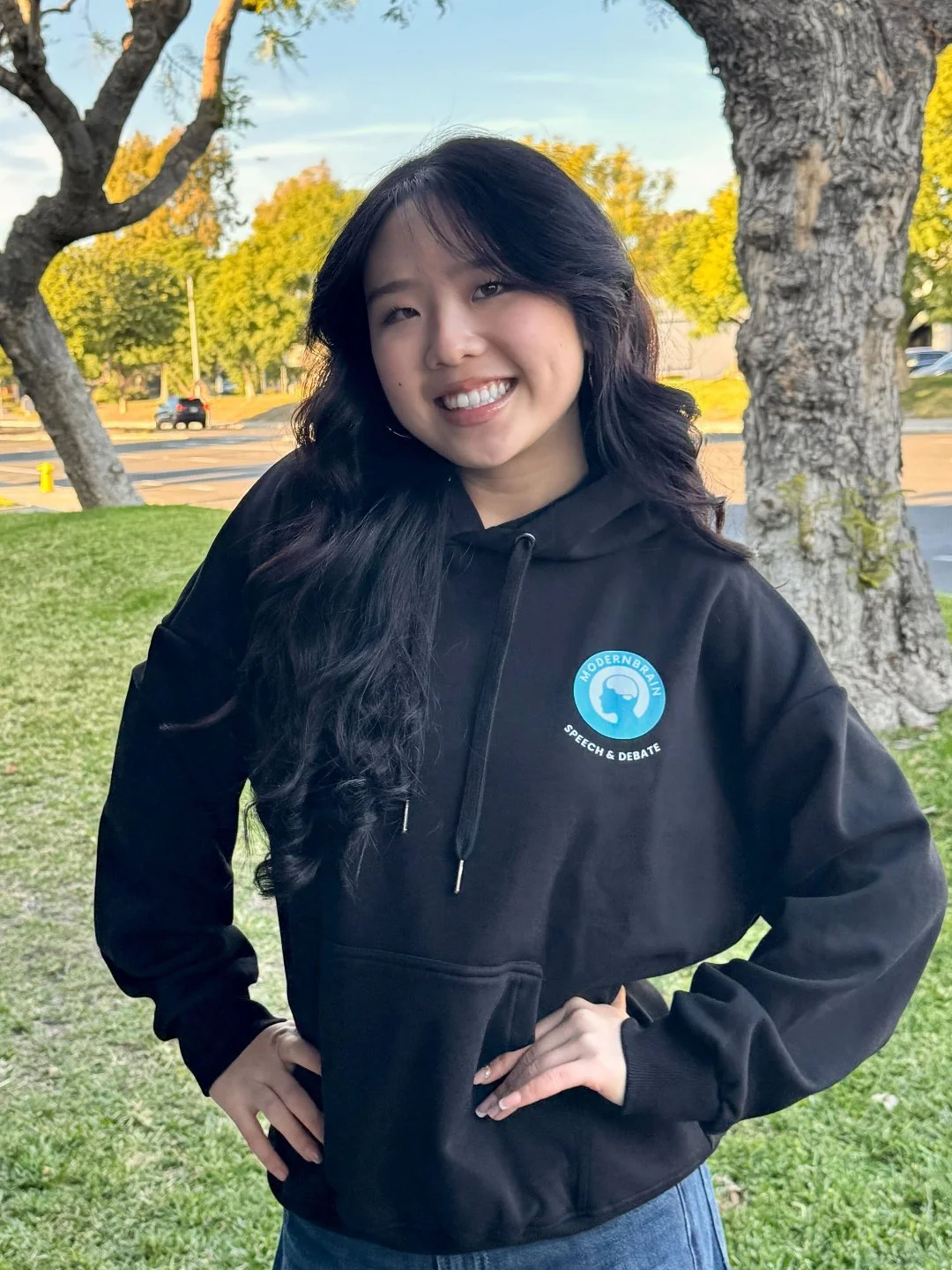 A young woman with long dark hair, smiling and wearing a black hoodie with a logo that reads 'Modern Brain Speech & Debate,' standing outdoors on grass with trees and a parking lot in the background.