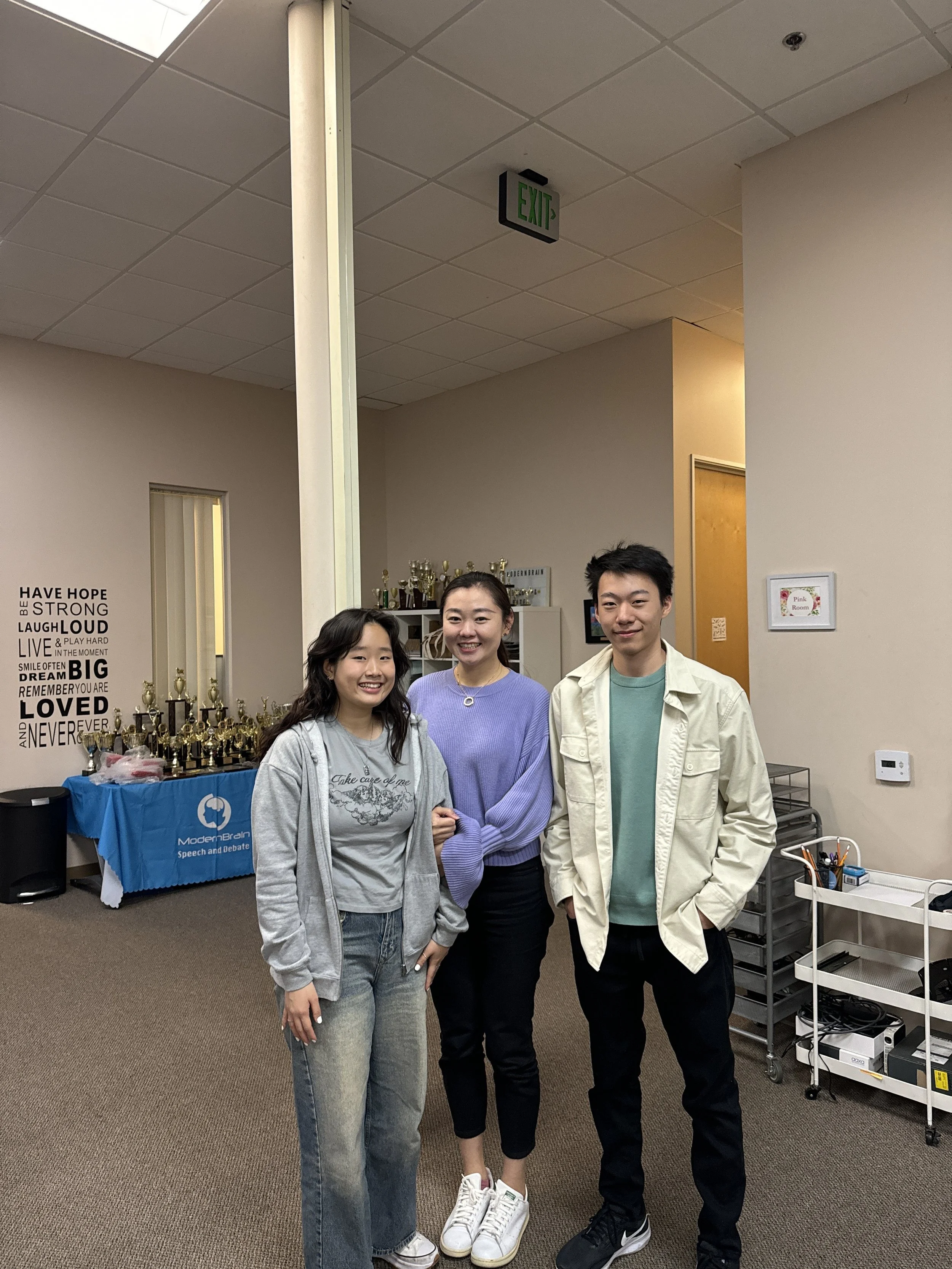 Three young people standing side by side in an indoor setting, smiling and posing for the camera, with trophies and motivational words on the wall behind them.