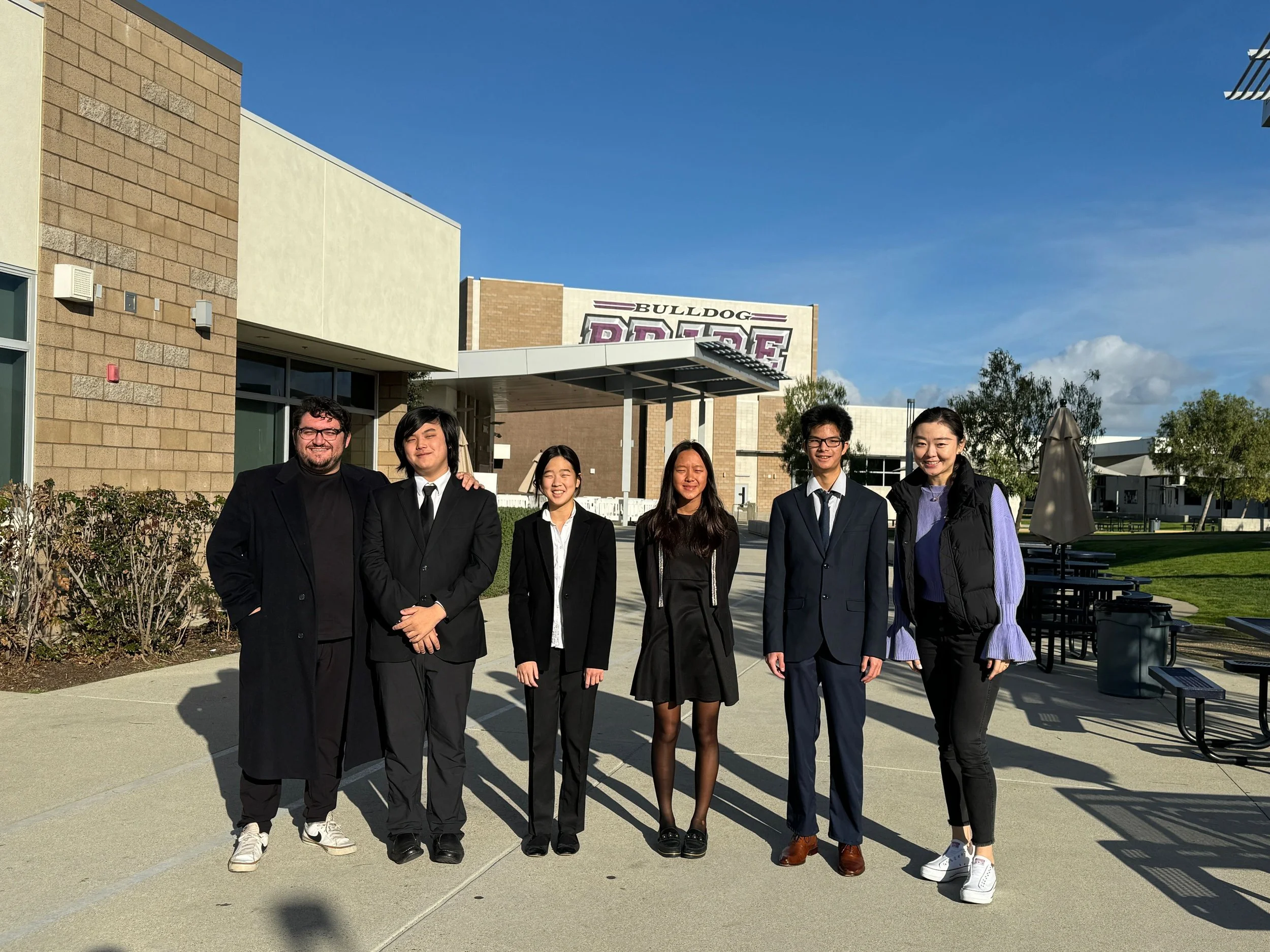 Group of six diverse young adults standing together outside on a sunny day, in front of a building with a sign that reads 'Bulldog' and 'Dons', dressed in semi-formal clothing, smiling at the camera.