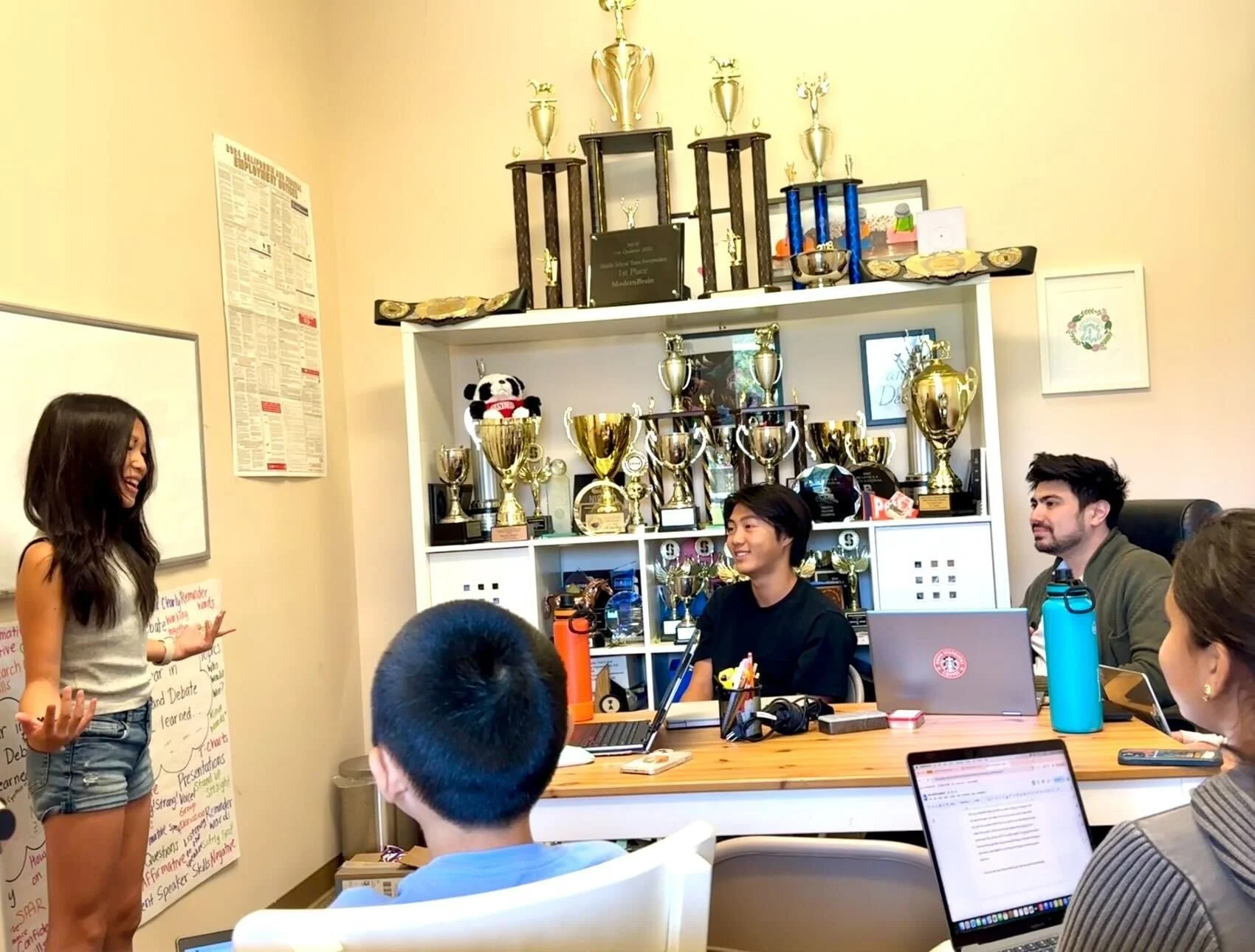 A young woman is presenting to a group in a room decorated with many trophies on shelves.