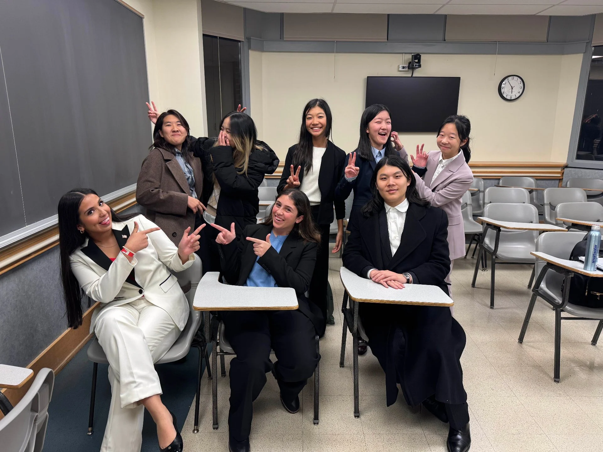 Group of nine young women in a classroom, some sitting and some standing, smiling and making peace signs, with school desks and chairs around them.