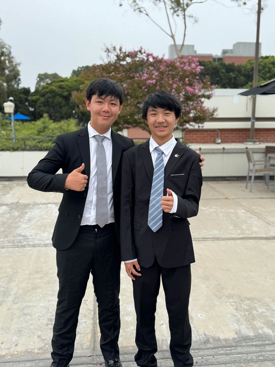 Two young men in suits standing outdoors, smiling, with pink flowering trees and buildings in the background.