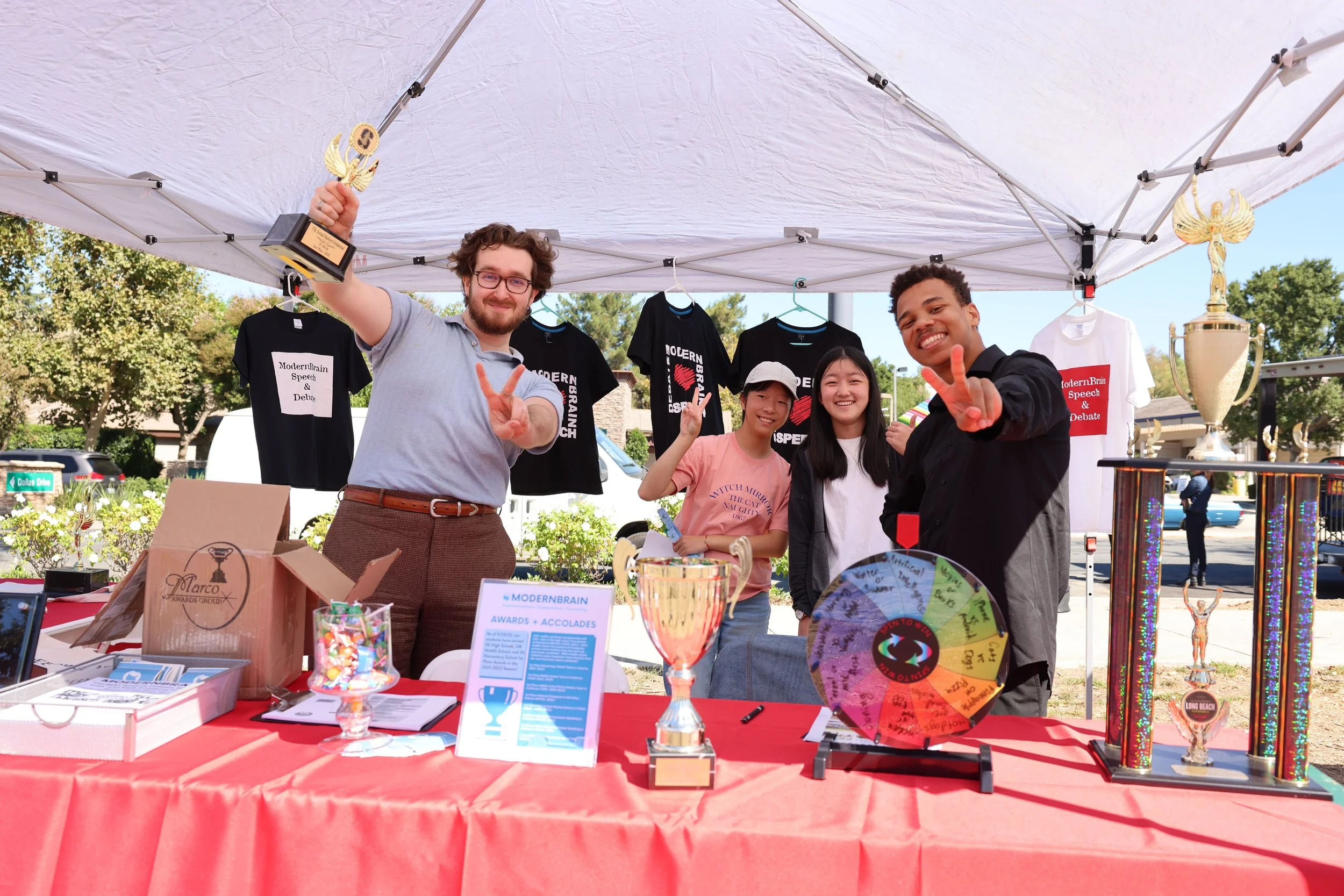 Four people smiling and holding trophies at an outdoor event booth with awards, shirts, and a prize wheel under a white canopy.