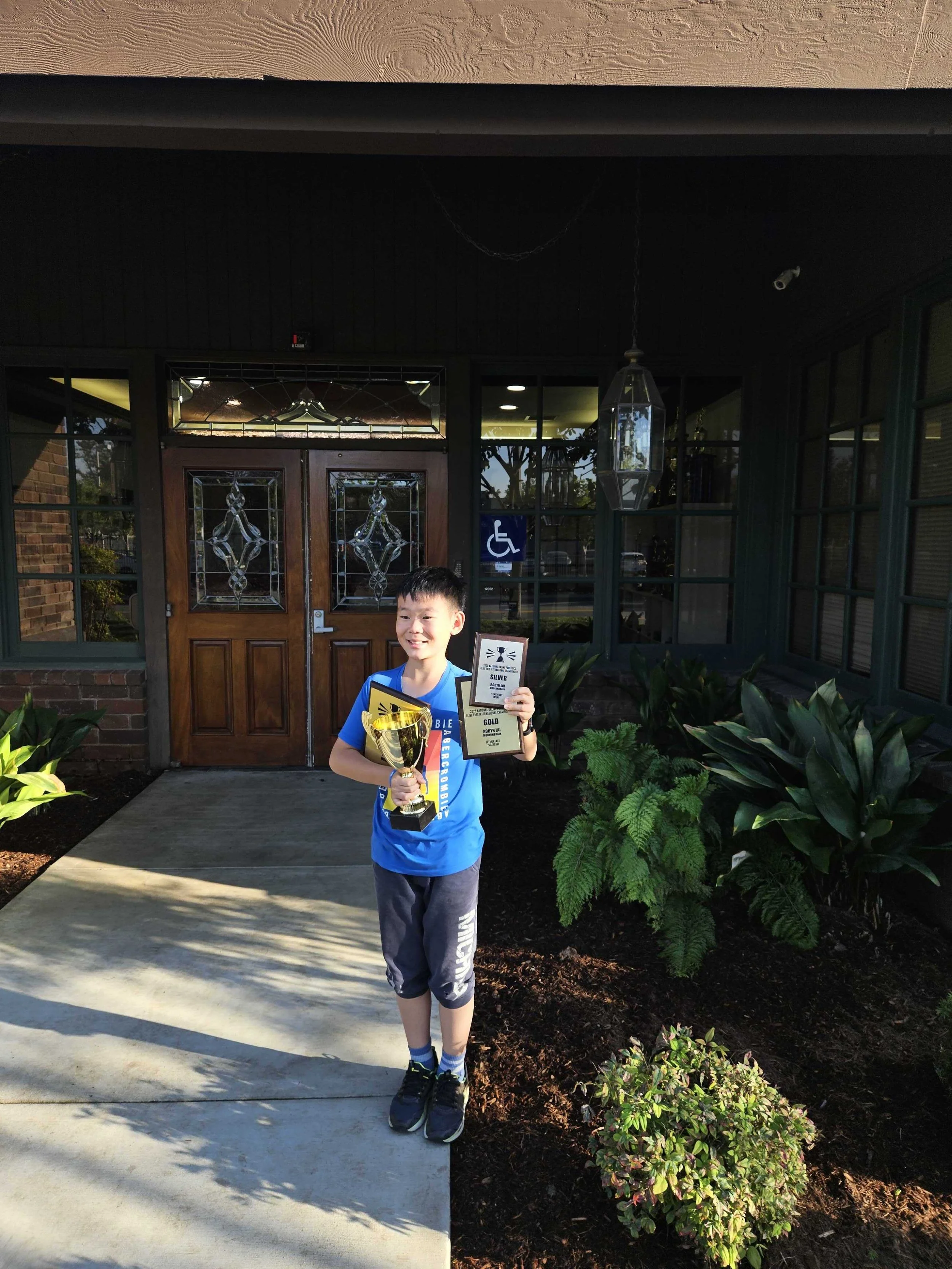 A young boy standing outside a building, holding a trophy and an award plaque, smiling after a competition or achievement.
