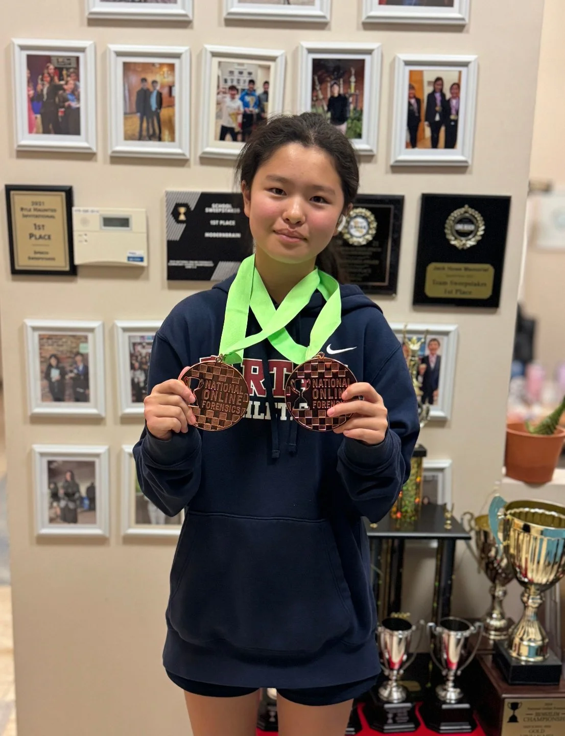 Young girl holding two bronze medals from a national online forensics competition in front of a display wall with framed photos and awards. The medals have the text 'NATIONAL ONLINE FORENSICS' on them.