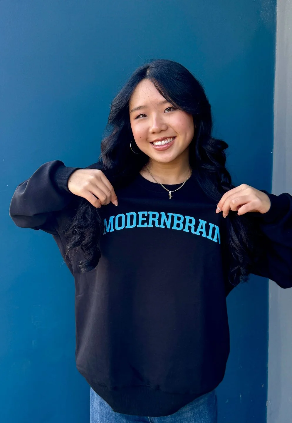 Smiling young woman with black hair and hoop earrings holding the collar of her black sweatshirt that reads 'MODERNBRAIN' in blue text, standing against a dark blue wall.