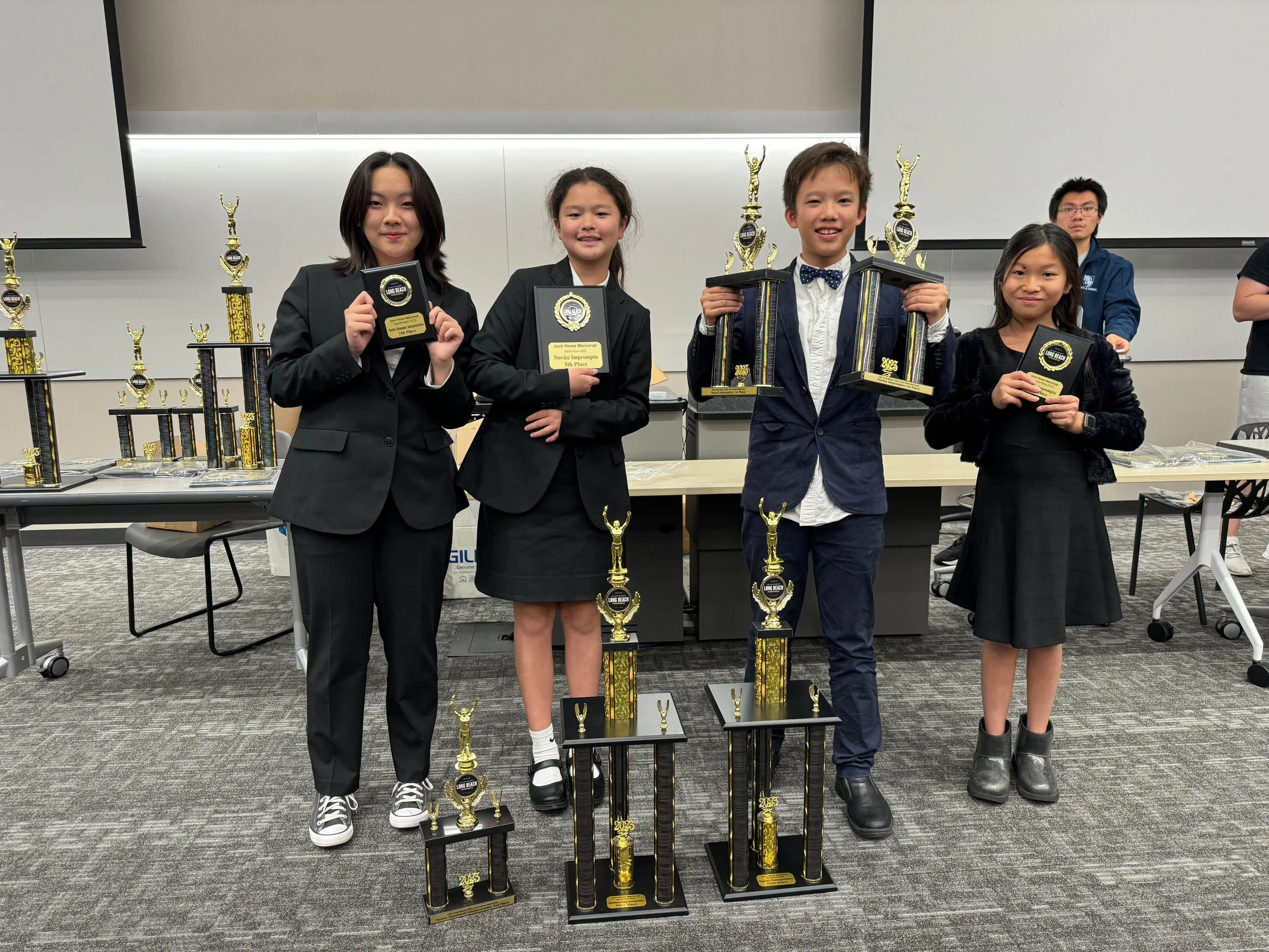 Four children standing in front of a table with multiple trophies, holding awards, celebrating a competition victory in an indoor setting.