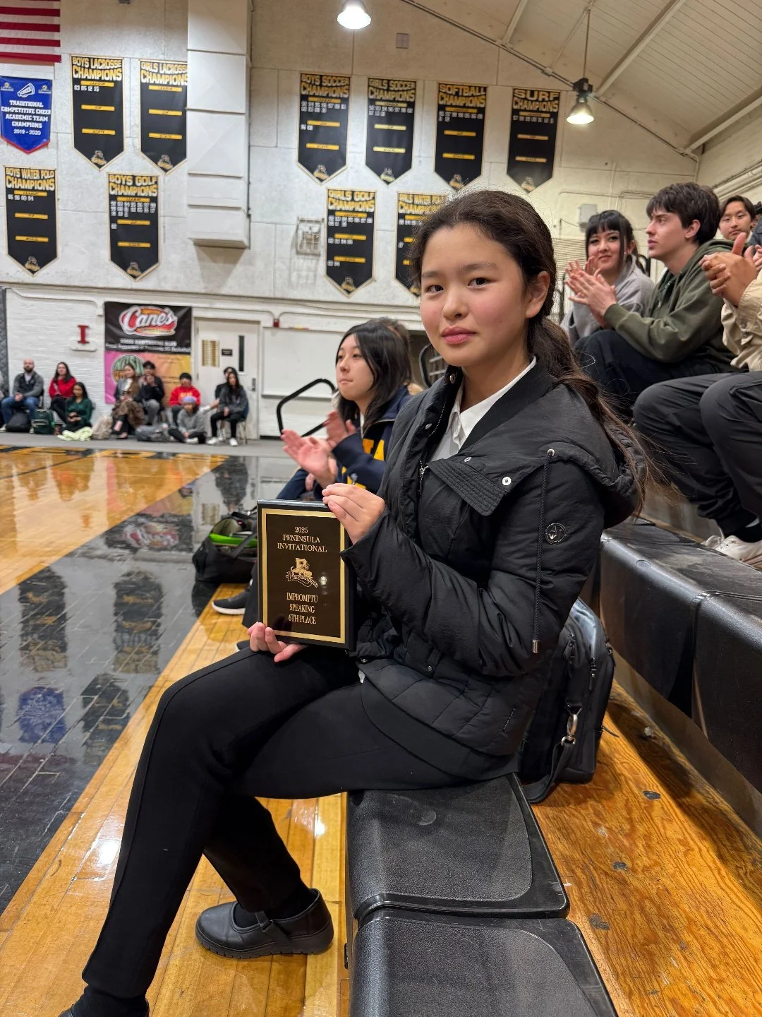 Young girl sitting on a gym bench holding a plaque that reads "2023 Peninsula Invitational, Promptly Speaking, 6th Place," in a gymnasium with banners on the wall and other people seated around, some clapping.