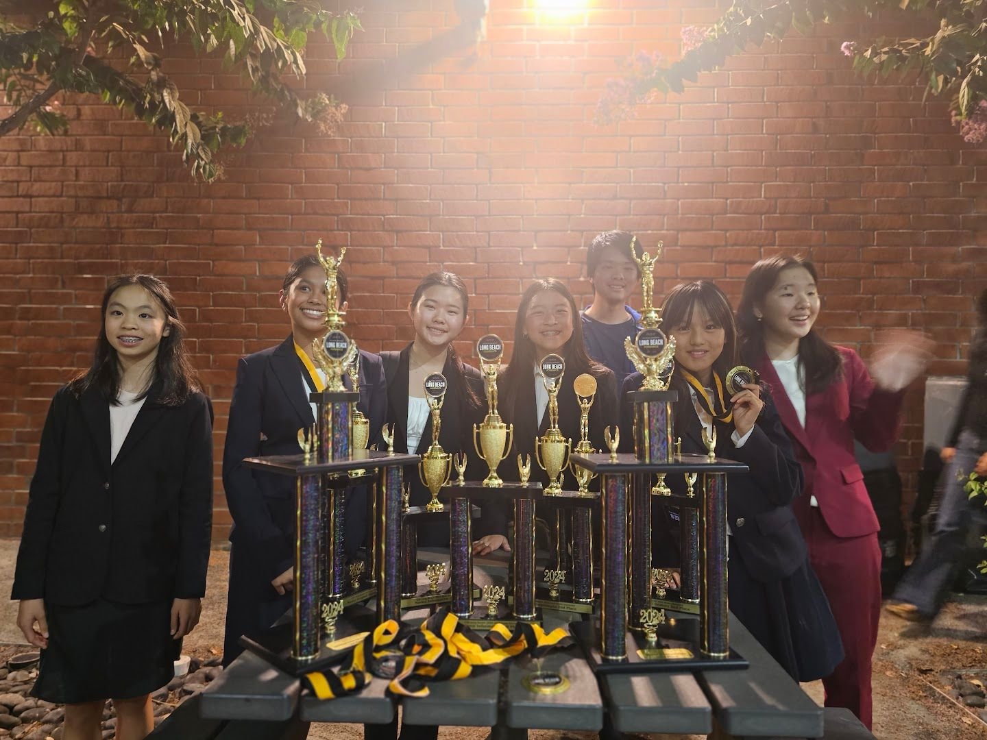 Group of seven young girls dressed in formal attire, standing behind a table with multiple trophies and medals, celebrating an achievement outside at night with a brick wall and trees in the background.