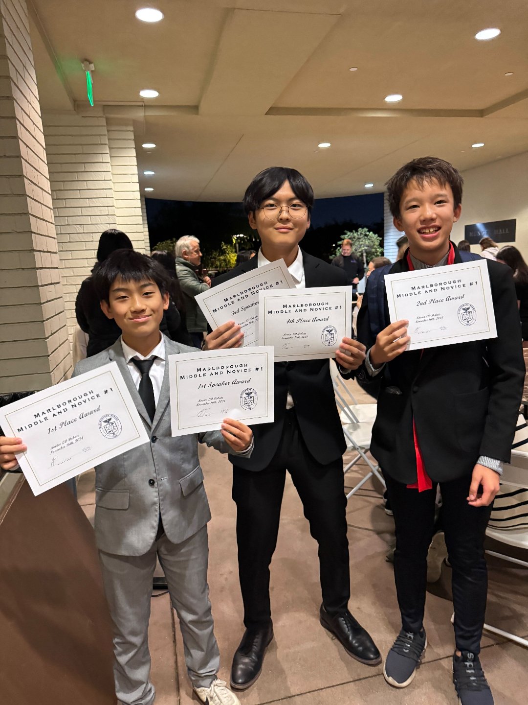 Three young boys in suits holding certificates at an awards ceremony, with other people in the background.