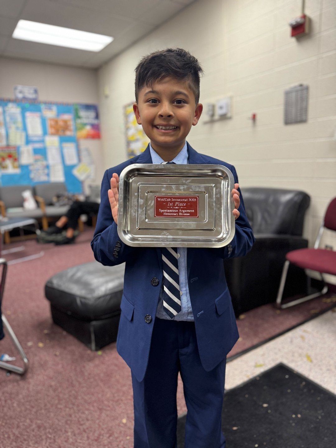 A smiling young boy in a blue suit holding a silver tray with a red plaque, standing in a room with chairs and bulletin boards.