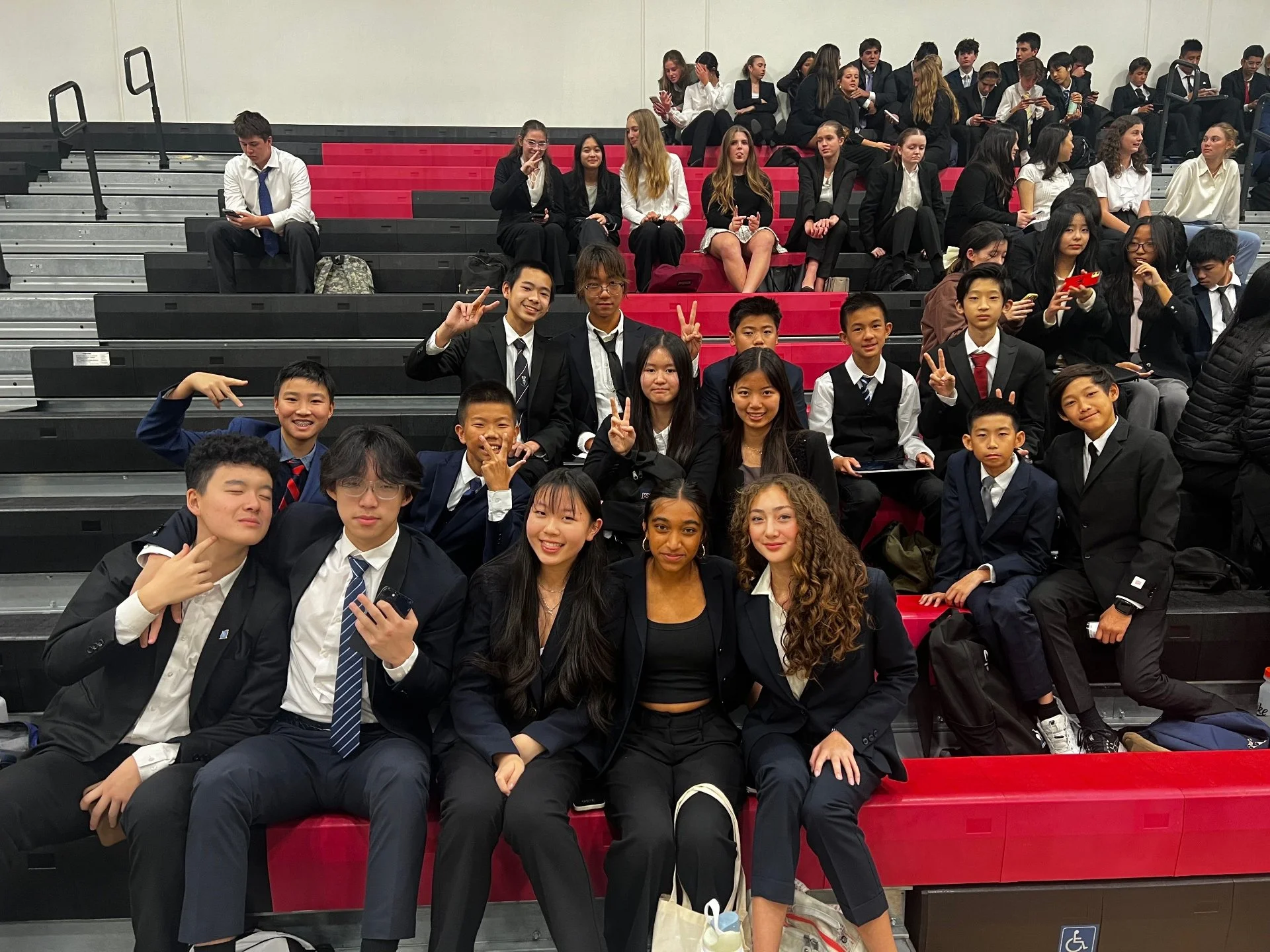 Group of students and teachers in formal attire sitting on bleachers in a school auditorium.
