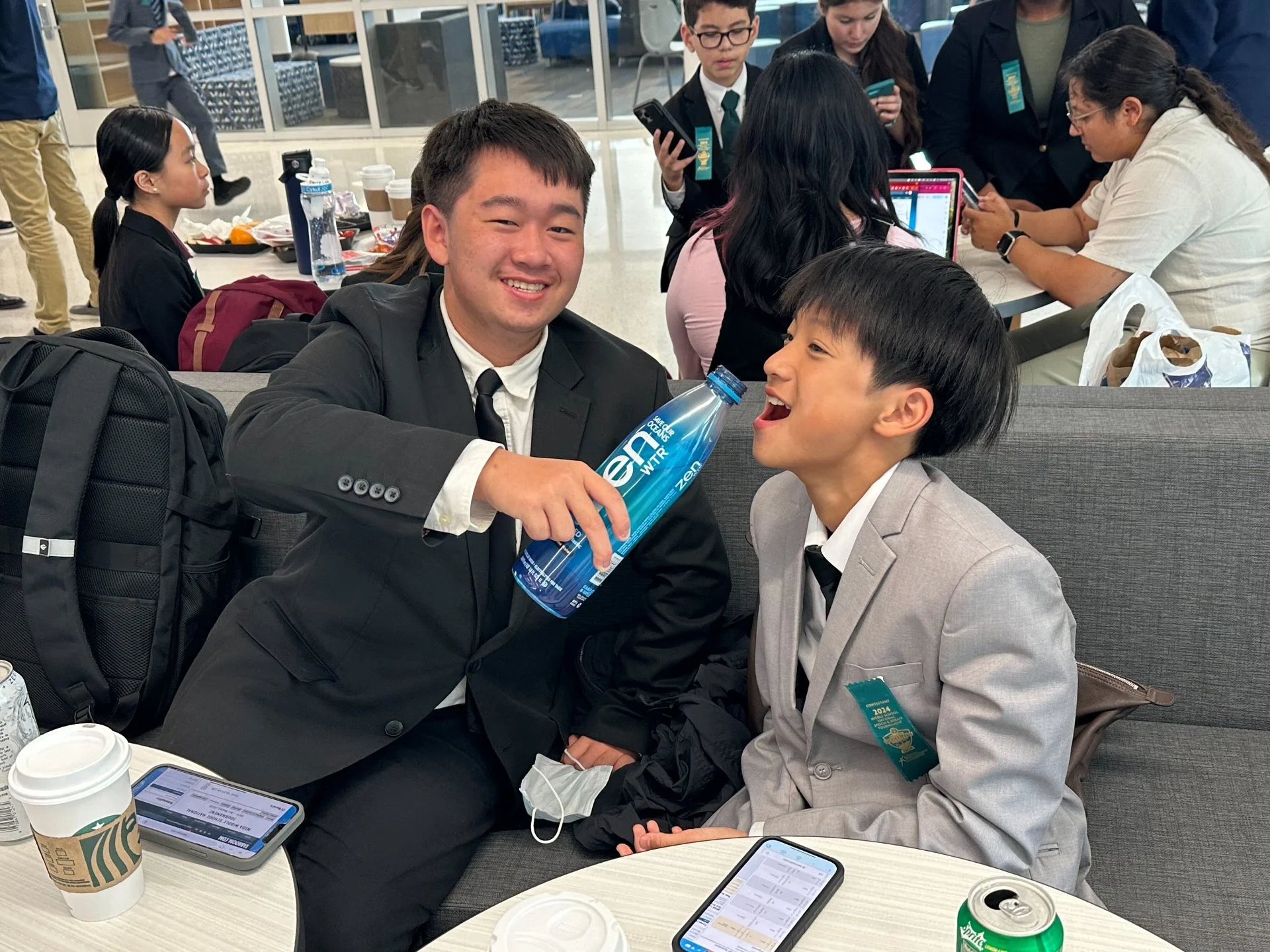 Two young men in suits, one holding a water bottle and the other smiling with an open mouth, sitting at a table in what appears to be a conference or meeting room with other people working and socializing in the background.