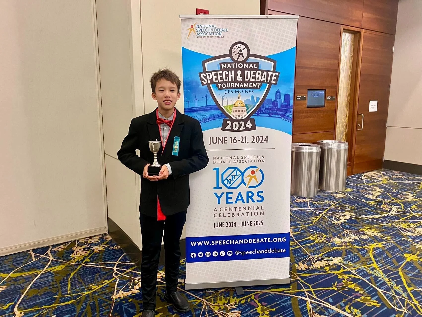 A young boy in formal attire holding a trophy standing next to a sign for the 2024 National Speech & Debate Tournament in Des Moines, celebrating the event's centennial anniversary.