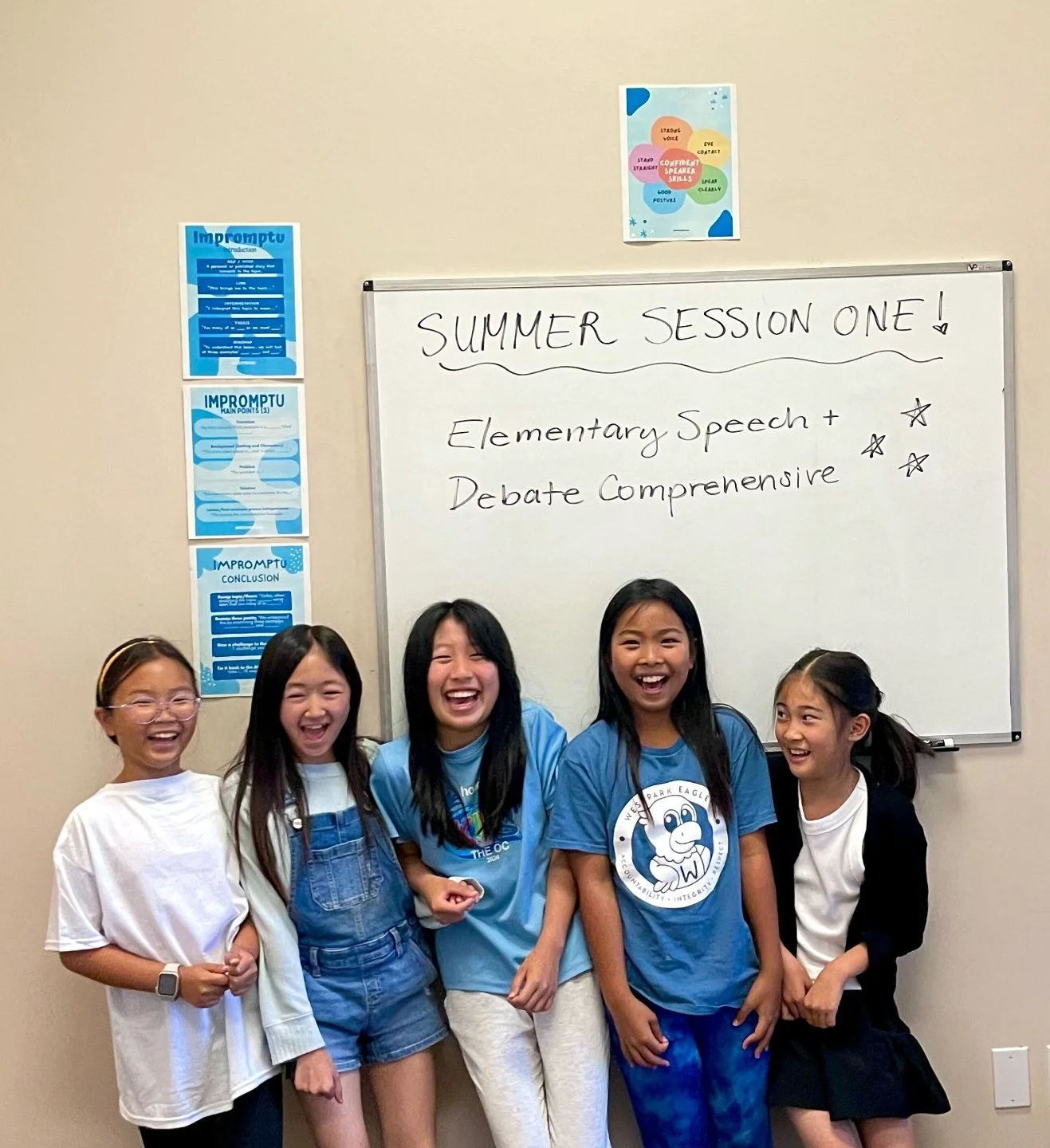 Five young girls smiling and laughing in front of a whiteboard with the text "Summer Session One! Elementary Speech + Debate Comprehensive." The girls are wearing casual clothes, and there are posters on the wall beside the whiteboard.