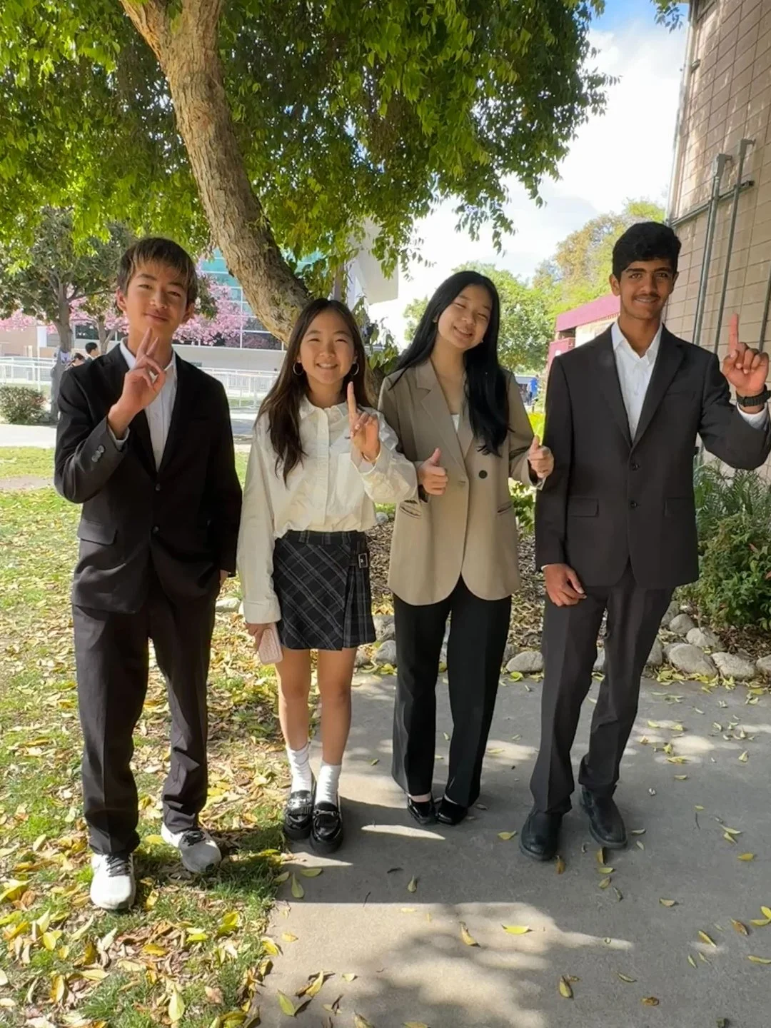 Four young students, two males and two females, dressed in formal attire posing outdoors under a tree, smiling and making peace signs and a thumbs-up gesture.