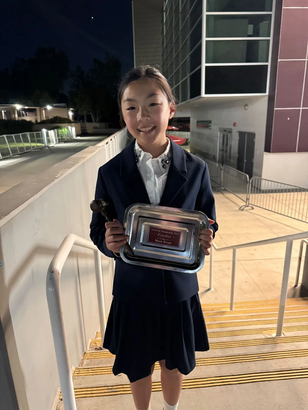 Young girl in school uniform holding a plaque and gavel, standing on school stairs at night, smiling.