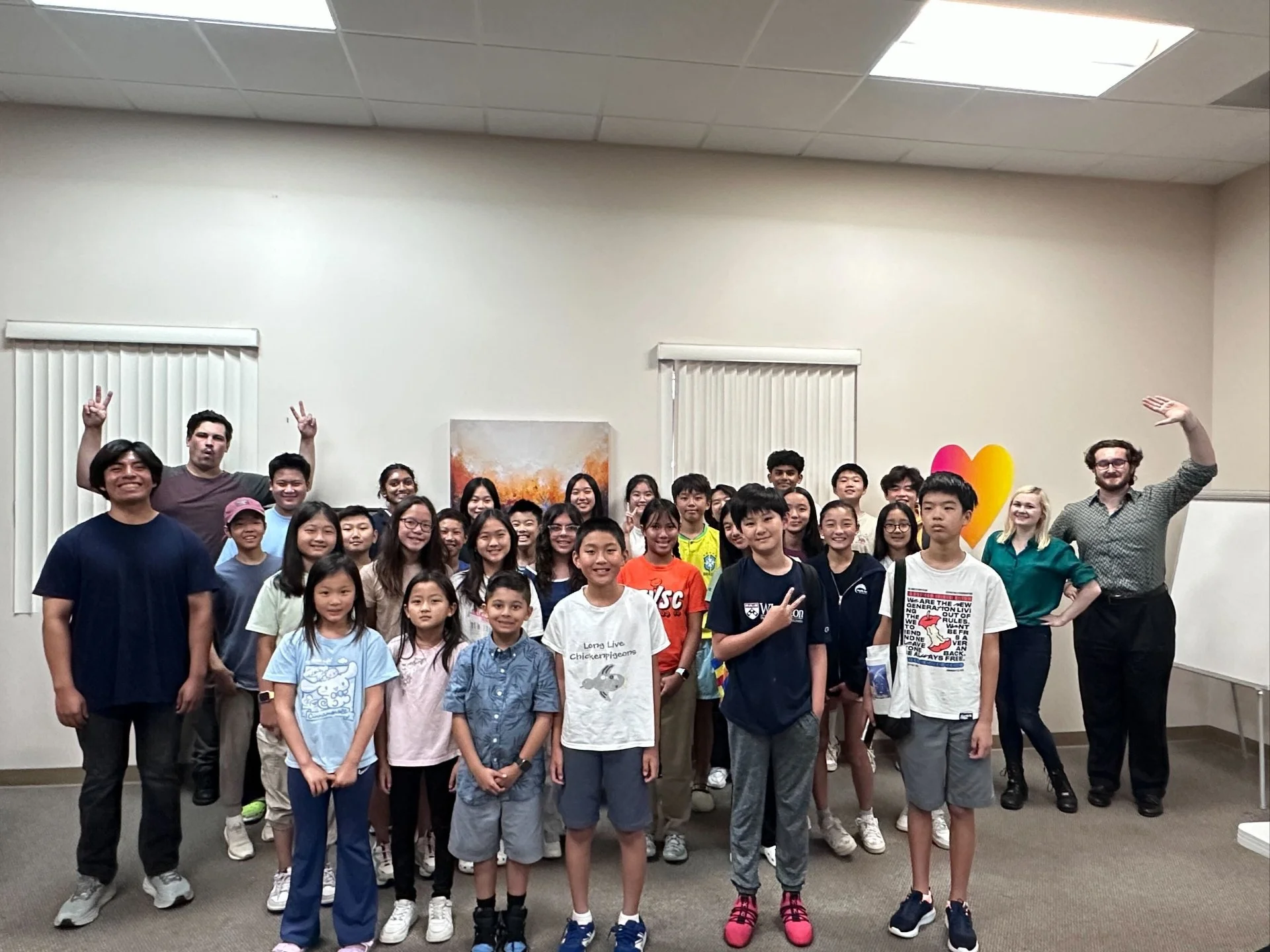Group of children and two adults standing in a room with painted wall art and white blinds.