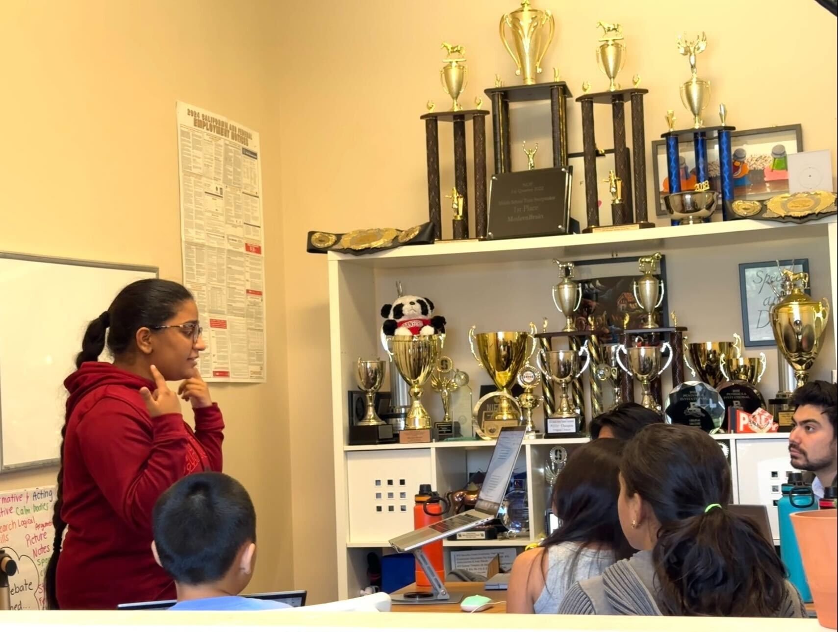 A woman in a red hoodie is speaking to a group of students in a classroom, with trophies and awards displayed on the bookshelf behind her.