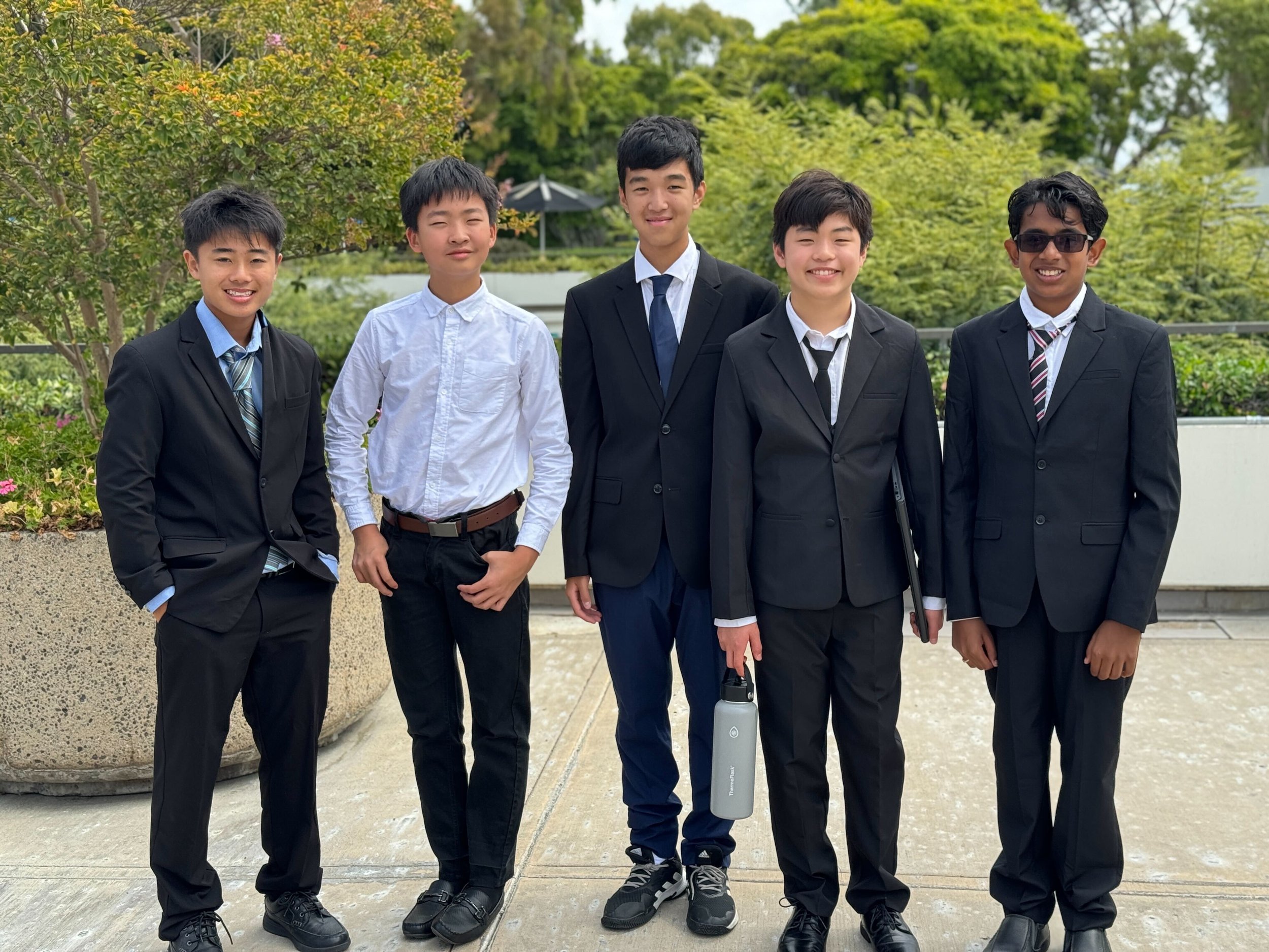 Five young men dressed in suits and business attire standing outdoors on a concrete patio with green trees and plants in the background.