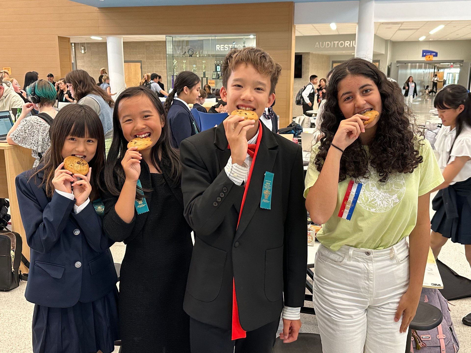 Four children smiling and eating cookies at a school event in a busy hall with other students in the background.