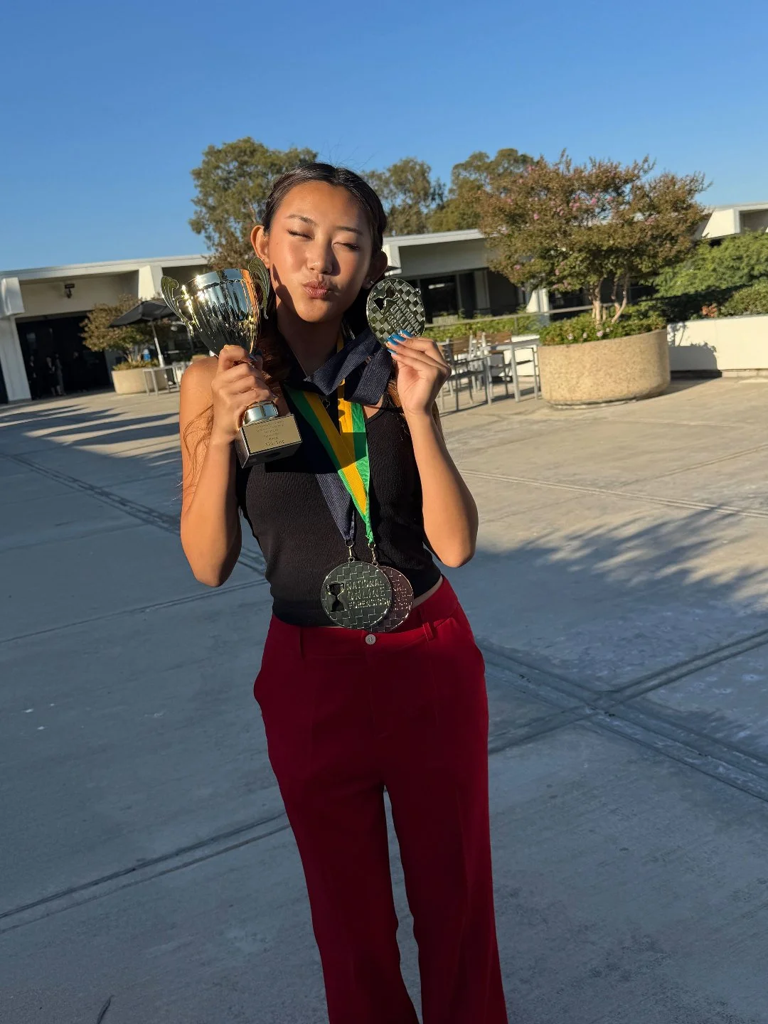 A young woman with a medal around her neck holds a trophy and a medal, standing outdoors in sunlight with modern buildings and trees in the background.